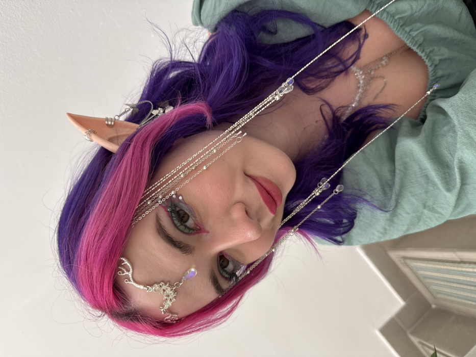 A woman with purple and pink hair, and pointed elf ears is looking at the camera. She is wearing a light green blouse and silver jewelry around her face. The jewelry includes chains with beads, a forehead piece with a purple gem, and embellishments around her eyes. She has green eyes with pink eyeliner. The background is a white wall and a partially visible window.