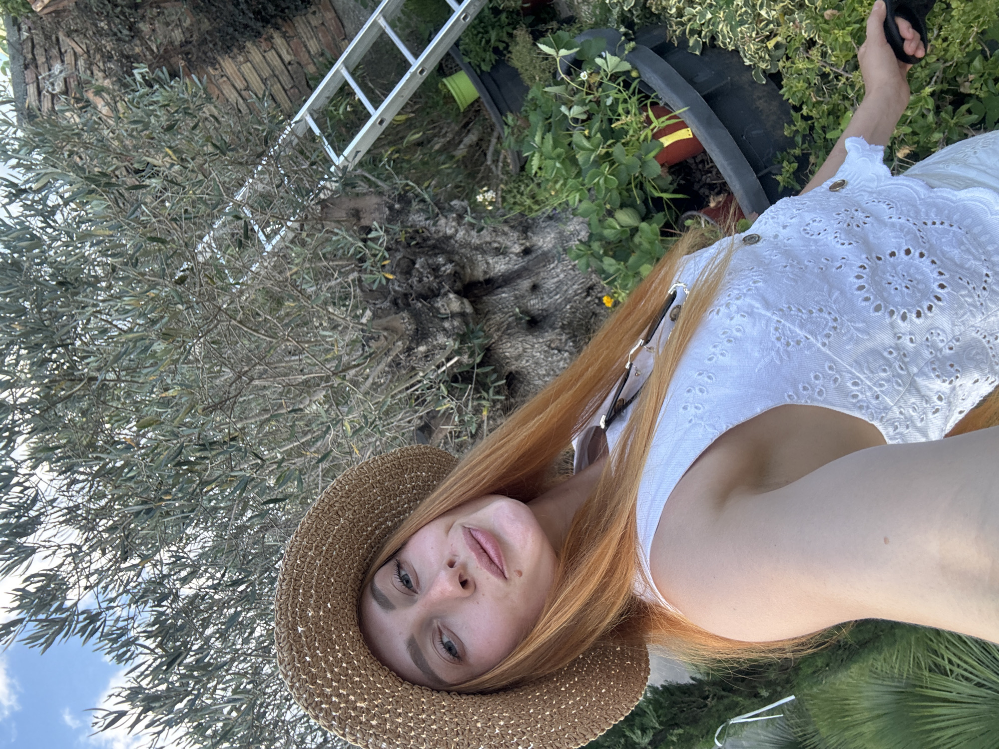 A woman with long, ginger hair and a straw hat is taking a selfie outdoors in a garden. She is wearing a white, eyelet top and is holding a black leash in her right hand. Behind her, there is a large, gnarled olive tree, a silver ladder leaning against a brick wall, and several potted plants, including strawberries. The scene appears to be located in Radanovići, Montenegro. The sky is blue with some clouds.