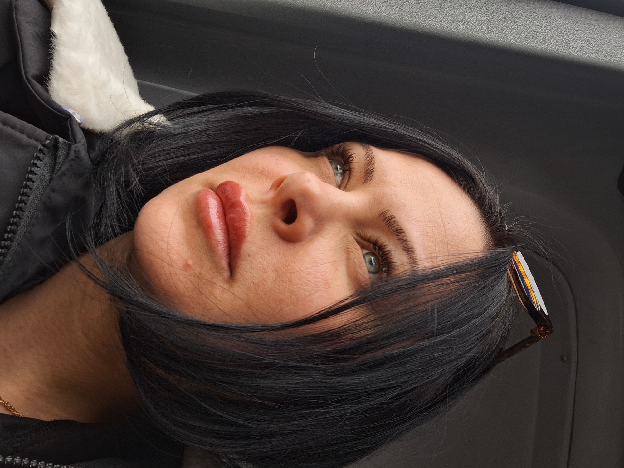 A close-up, slightly upward-angled shot features a woman with black hair and light eyes. She is wearing a dark jacket with a white fur lining visible at the collar. A pair of tortoiseshell sunglasses are perched on the side of her head. Her lips are prominent and appear to have a glossy finish. A thin gold chain is visible at her neck. The background is a neutral gray, possibly an interior car setting. The image does not provide sufficient information to determine a location in Moscow, Russia.