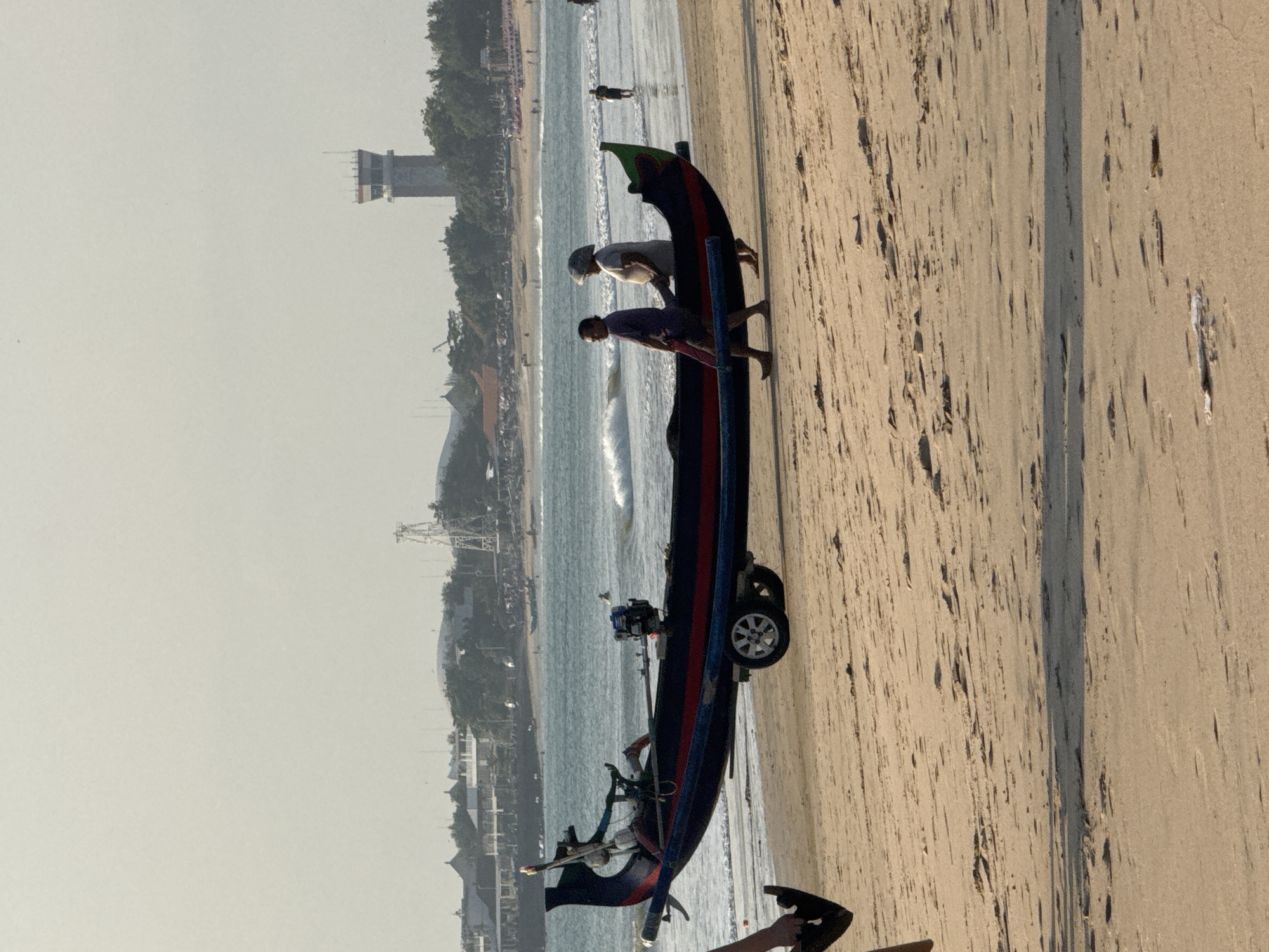 This image captures a beach scene in Kuta Selatan, Indonesia, with a long, narrow boat on a trailer positioned on the sand near the water. The boat is dark blue with red and black stripes. Two individuals are walking beside the boat, one in a purple shirt and dark shorts, and the other in a light-colored shirt and a hat. Further out, the ocean has moderate waves, and a few people are in the water. In the background, a townscape with buildings and trees is visible, along with an air traffic control tower. The sky is overcast and hazy.