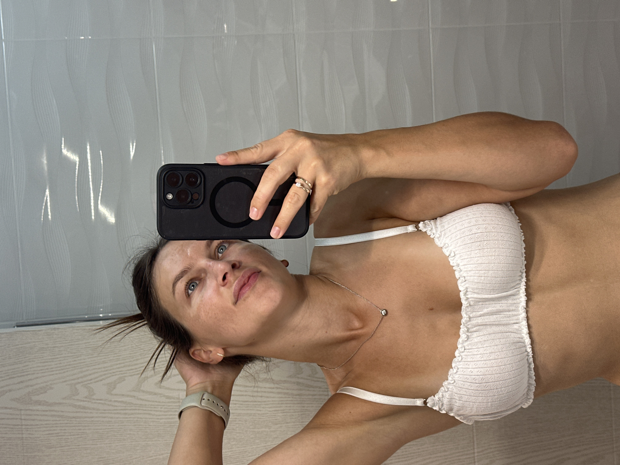 A woman takes a mirror selfie in a bathroom. She is wearing a white bralette and has a face mask on. Her hand is raised to her head, and she is holding a black smartphone. The bathroom walls are tiled in a wave pattern. The woman appears to be in a private residence, likely a home in Bečići, Montenegro.