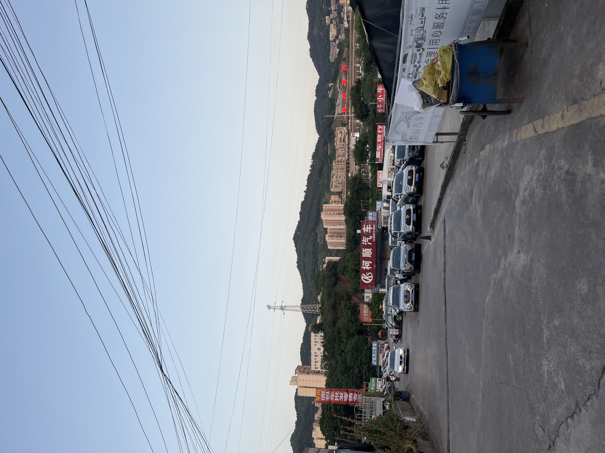The image depicts a street scene in the city of Banan, China, showcasing a row of parked cars on the side of the road with a concrete sidewalk in the foreground. The backdrop features a hillside dotted with buildings and trees, while overhead power lines intersect the sky. Chinese characters are visible on banners and signs along the street, indicating local businesses. A blue cart is visible on the sidewalk, suggesting commercial activity.