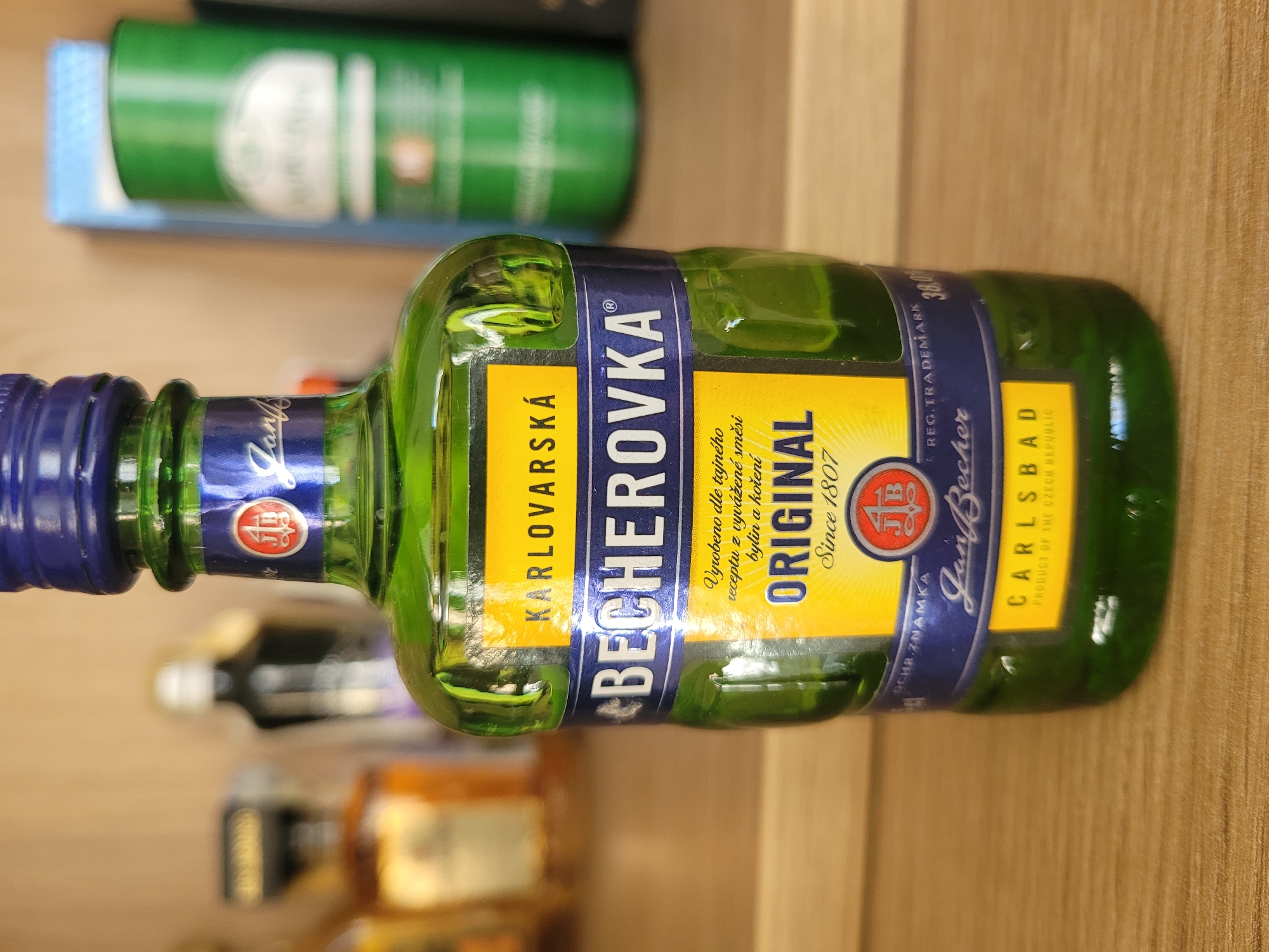 The image shows a close-up of a green glass bottle of Becherovka Original, a liqueur. The bottle has a blue cap and several labels. The main label is yellow with blue text that reads "BECHEROVKA ORIGINAL". There is also a smaller yellow label at the bottom that reads "CARLSBAD PRODUCT OF THE CZECH REPUBLIC". The bottle is standing on a wooden surface, likely a shelf or table. In the background, there are out-of-focus bottles and shelves, suggesting it is part of a collection or display. The lighting is consistent and focused on the bottle, highlighting its details. The image does not provide any information about the location being in the city of Kyiv, Ukraine.