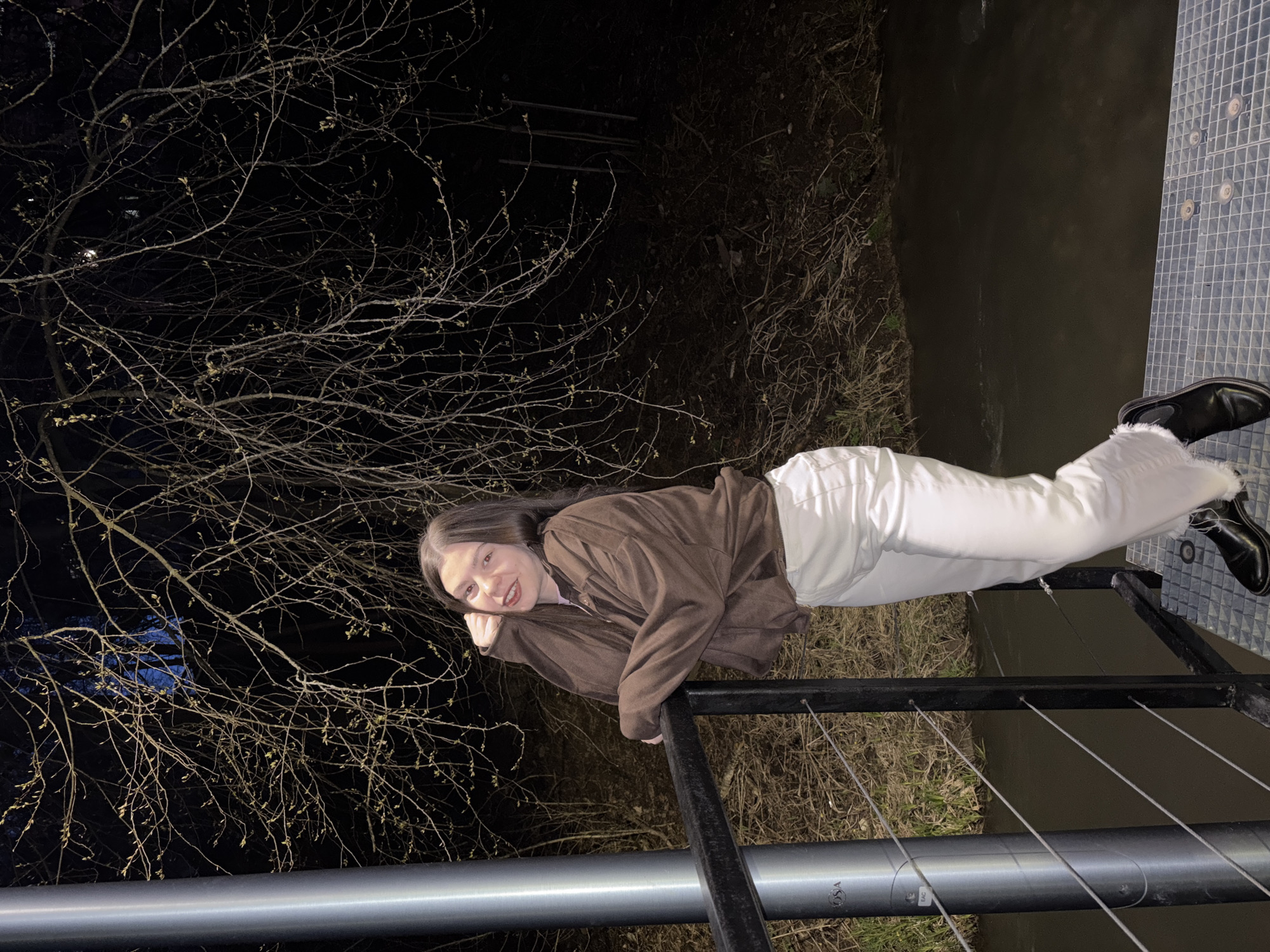 A young woman is pictured at night, leaning casually on a metal railing, looking towards the camera with a slight smile. She supports her head with her right hand, her elbow resting on the top bar of the railing. She has long, dark hair and is wearing a dark brown jacket over a lighter top, paired with white pants and dark shoes.
The scene is outdoors, likely beside a body of water visible to the right. The water's surface appears dark and still, reflecting minimal light. A metal grate platform is visible beneath her feet and extending to the right. In the background, against the dark night sky, are numerous bare tree branches, some with small buds, suggesting late winter or early spring. The lighting is artificial, illuminating the woman and some of the branches, creating a stark contrast with the dark background.
The railing she is leaning on consists of thick horizontal pipes and thinner vertical bars, with several tension wires running horizontally below the top bar. On one of the lower metal pipes, the letters "OSA" and "EAC" are visible. The ground beyond the railing to the left appears to be covered in dry grass and foliage. The image captures a moment in an urban or park setting in Perm, Russia, during the night.