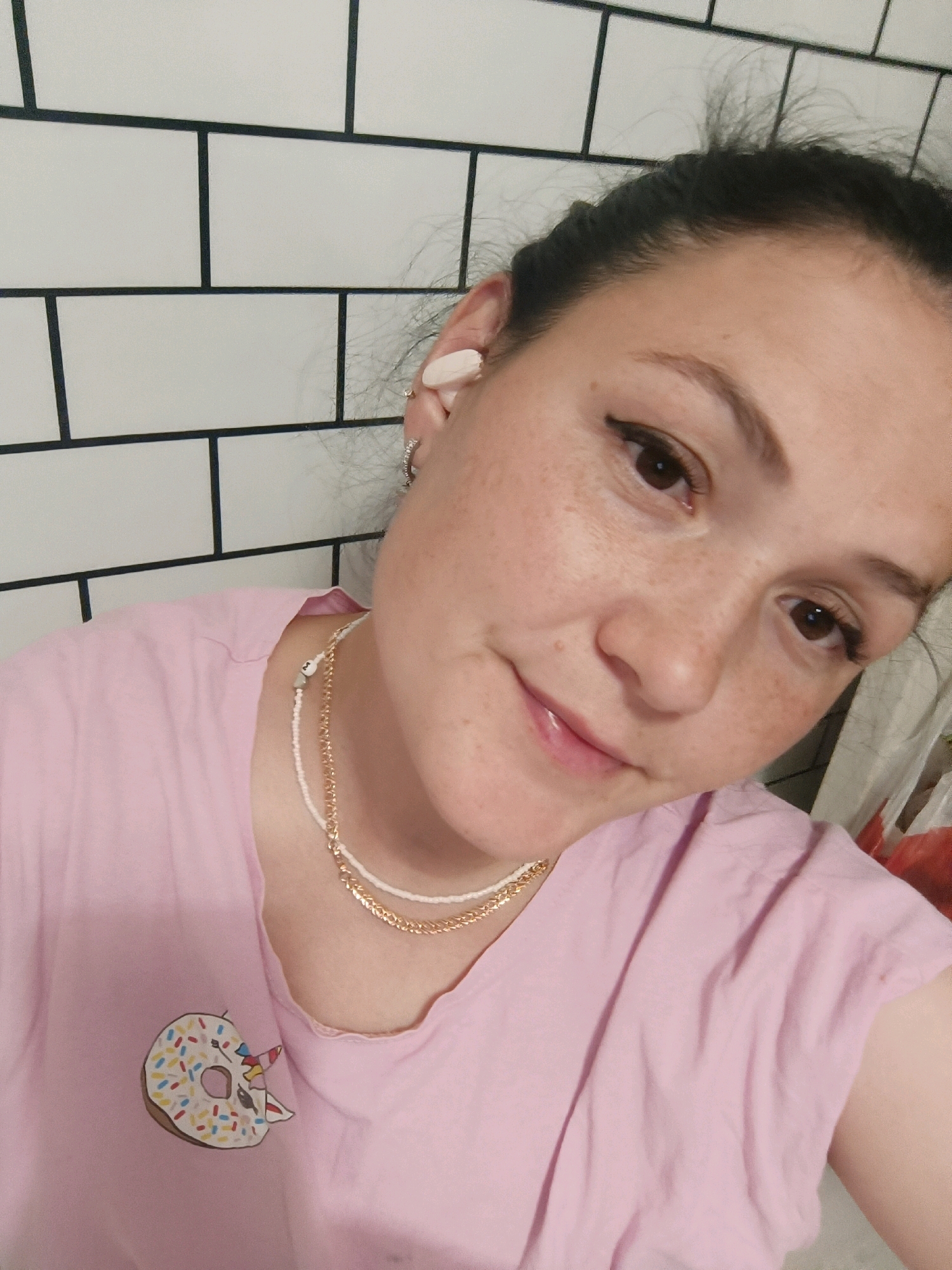 A woman is taking a selfie in a bathroom with white subway tiles. She is wearing a pink t-shirt with a donut design and two necklaces. She has on AirPods and several rings. Her hair is pulled back.