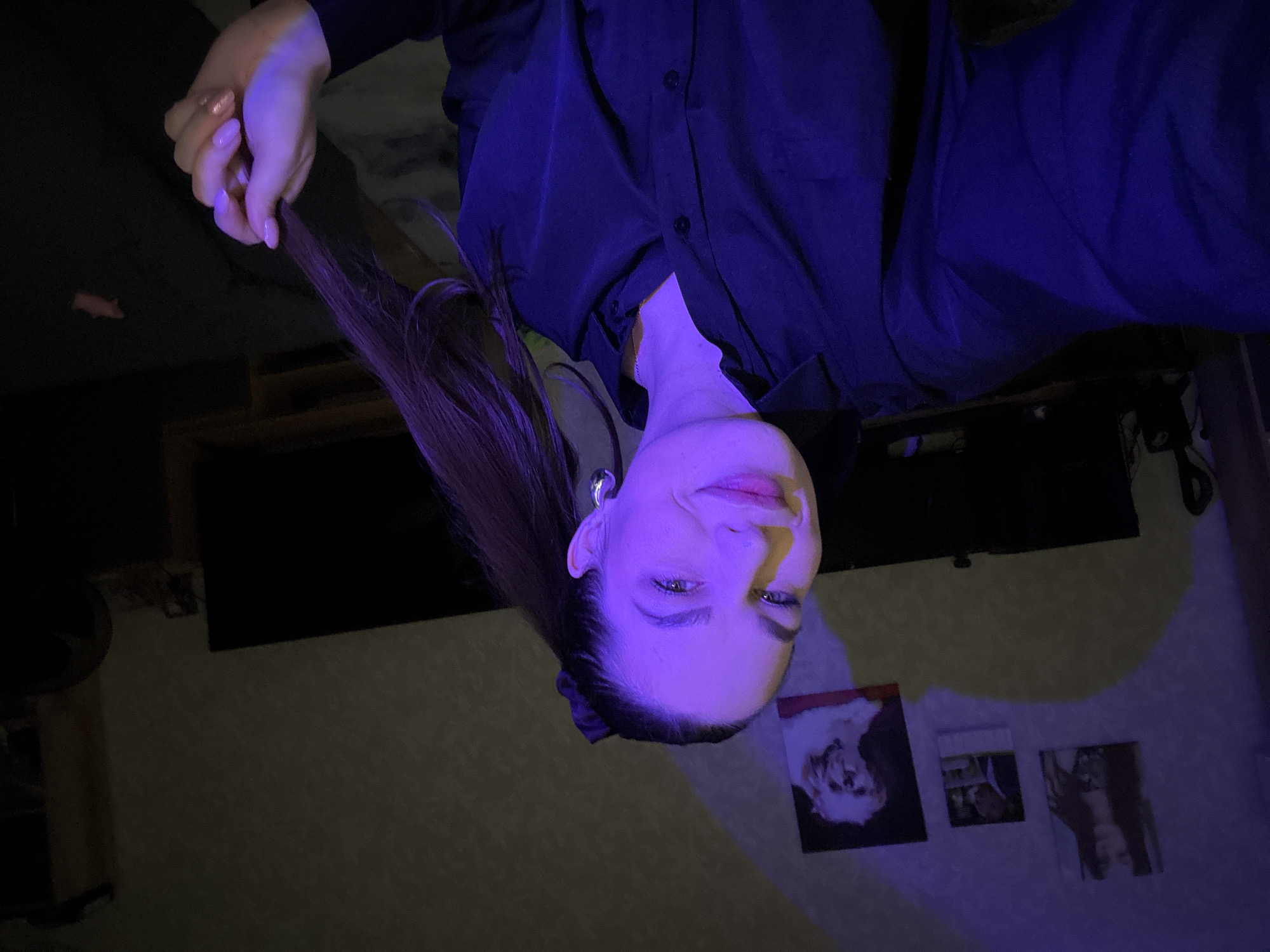 The image shows a young woman indoors, illuminated by purple light. She is wearing a black button-down shirt and has her hair in a ponytail. She is holding a strand of her hair in her right hand, and her nails are painted a light pink color with some glitter. In the background, there are several framed pictures on the wall, including one of a dog. There are also large dark screens, possibly televisions, and furniture. The overall impression is of a dimly lit room with the subject posing for a selfie. The context of Chaikovsky, Russia is not discernible from the image itself.