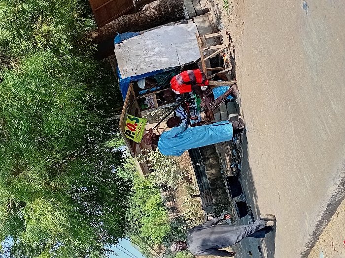 The image depicts a daytime street scene in Sharada, Nigeria. A paved road with a raised curb occupies the lower half of the frame. On the right, a makeshift vendor's stall, featuring a blue tarp roof, white side paneling, and a prominent yellow sign indicating "P.O.S Available," is positioned beneath a large, leafy green tree. Various small items are visible on shelves within the stall.
Four adult males are present. In the left midground, a man in a dark grey traditional tunic (kaftan) and cap walks away from the camera, heading left. Centered, a man in a light blue traditional tunic and cap stands with his back mostly to the camera, facing the stall. To his right, a man in a red t-shirt and dark trousers sits on a simple wooden stool, engaged with the stall. A fourth man, wearing a white cap, is partially visible behind the stall's counter, seemingly attending to customers. On the ground near the curb, two dark, round pots are visible amidst minor debris. The background shows sparse vegetation and faint structural elements under a clear sky.