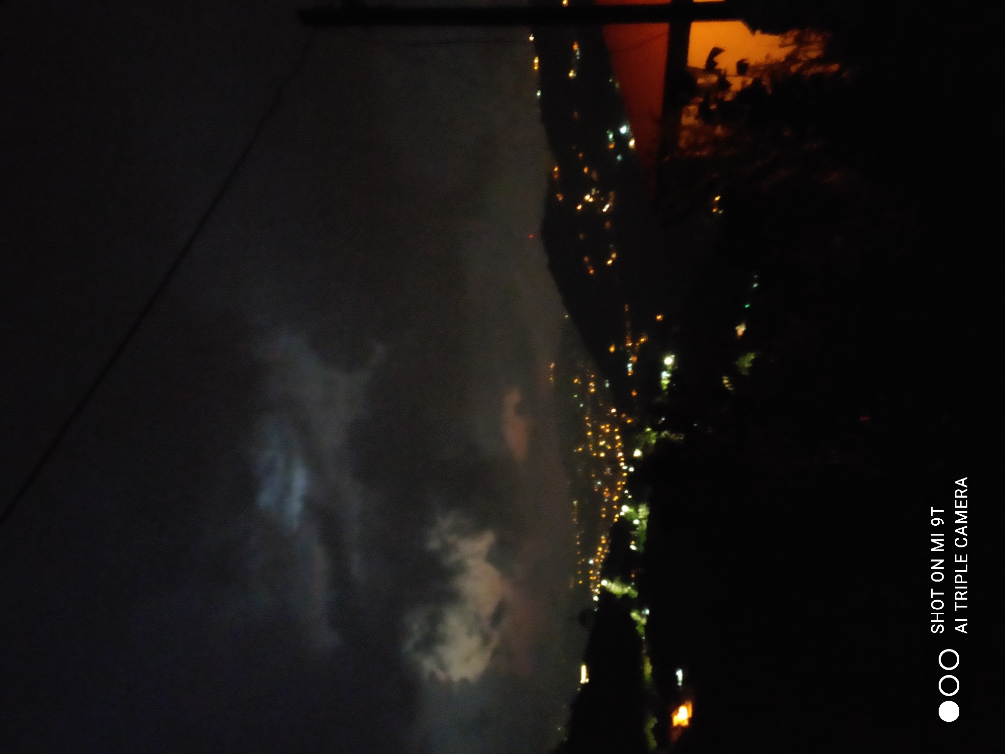 This is a nighttime, high-angle shot of a city, possibly Akçaabat, Turkey, viewed from a hillside. The sky is dark and cloudy, with some areas of lighter clouds and a hint of moonlight breaking through. Below, the city is illuminated by a multitude of bright, blurred lights, suggesting a dense urban area spread across hills. The foreground is dominated by the dark silhouettes of trees and a building with an orange-lit roof. The lower portion of the image displays a watermark: "SHOT ON MI 9T AI TRIPLE CAMERA." There are no discernible people or specific actions taking place, only the ambient glow of a city at night.