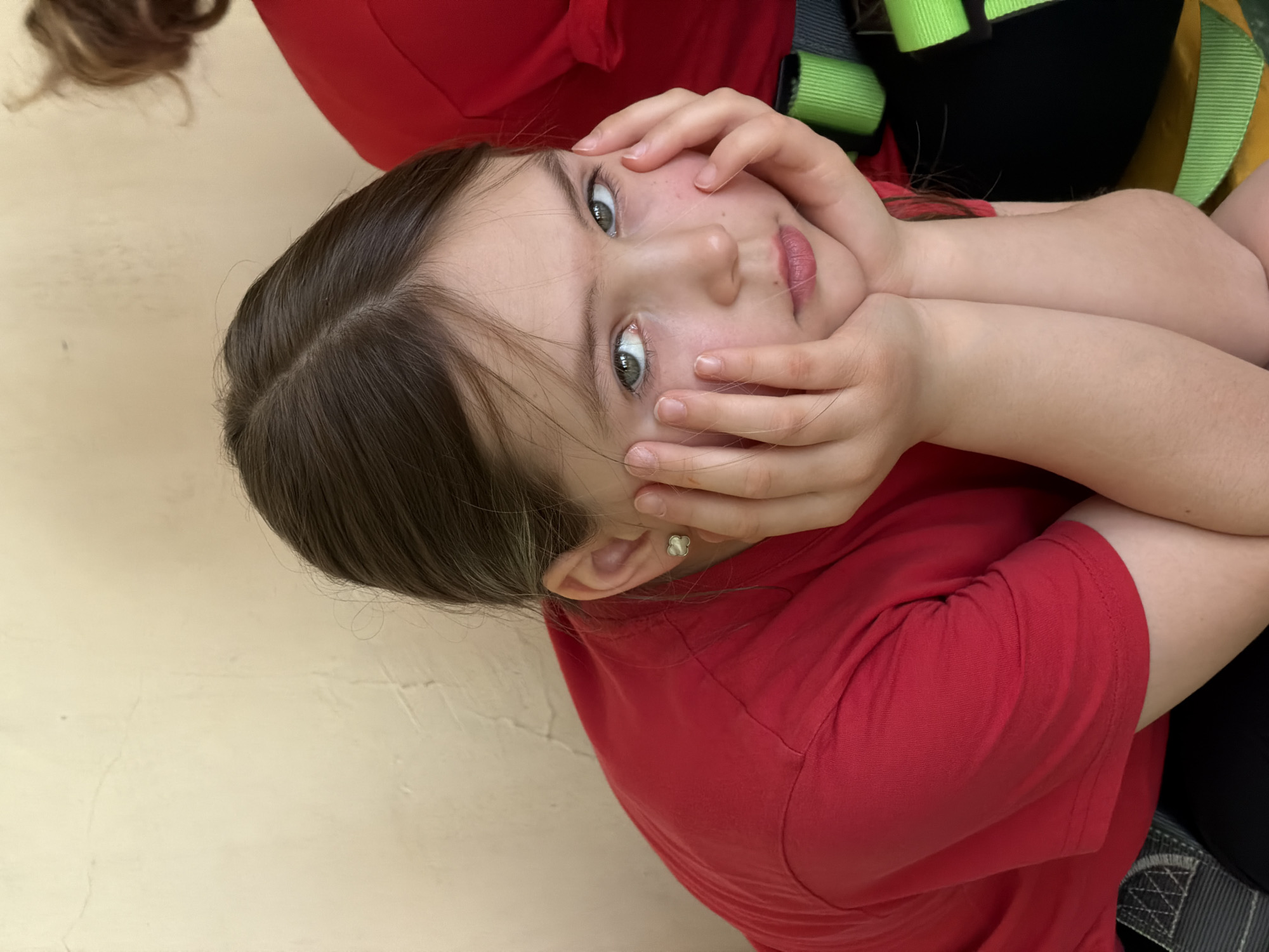 A young girl with light skin, brown hair, and green eyes poses for the camera. She is wearing a red shirt and resting her face in her hands. A small, flower-shaped earring is visible in her left ear. Another person wearing red and a safety harness is partially visible in the background. The location is unknown.
