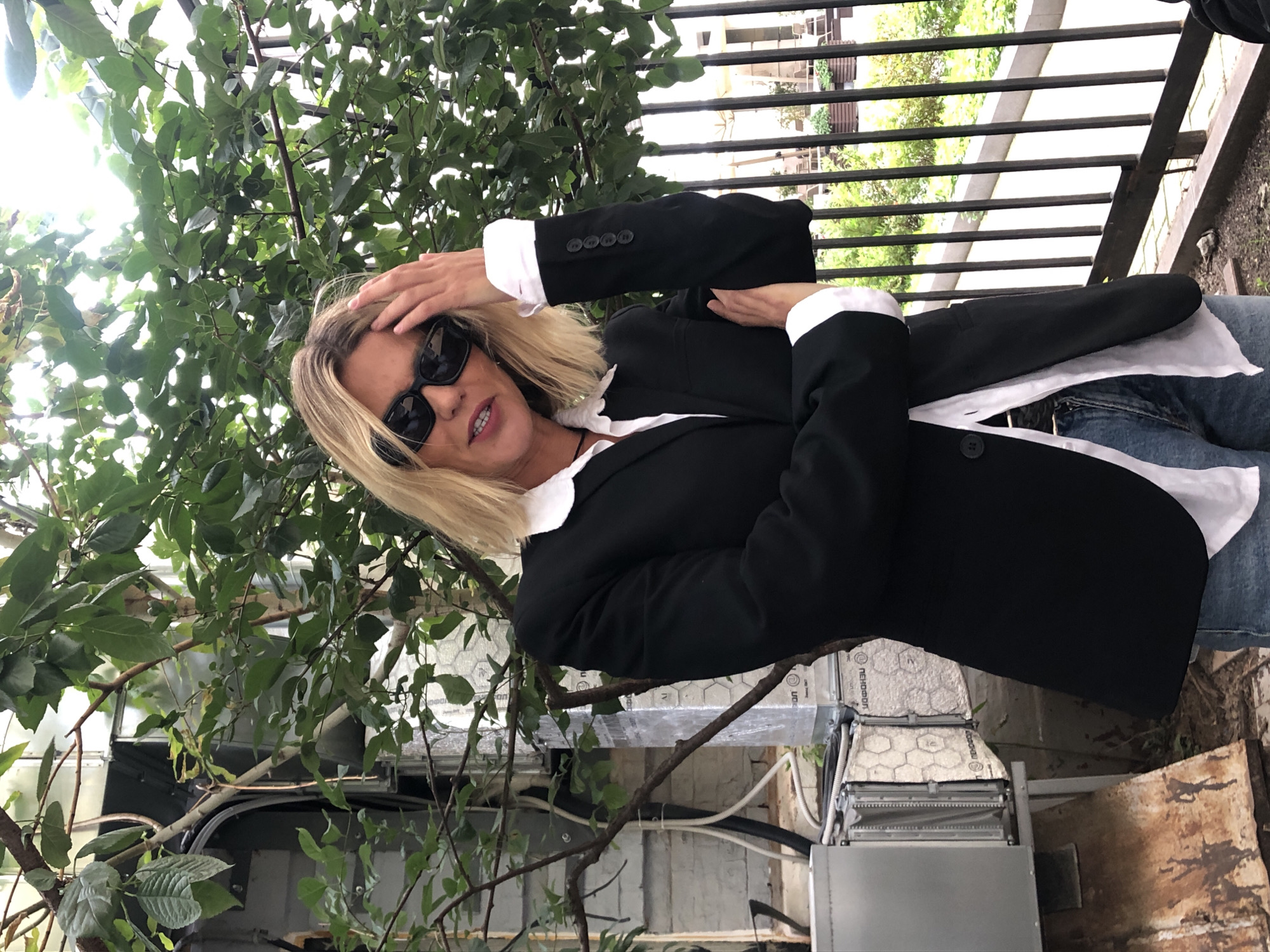 A blonde woman with sunglasses and a black blazer stands in front of a leafy green tree and a metal fence. She is wearing a white shirt and blue jeans. The background shows industrial equipment and urban greenery. The scene appears to be outdoors in an urban setting.