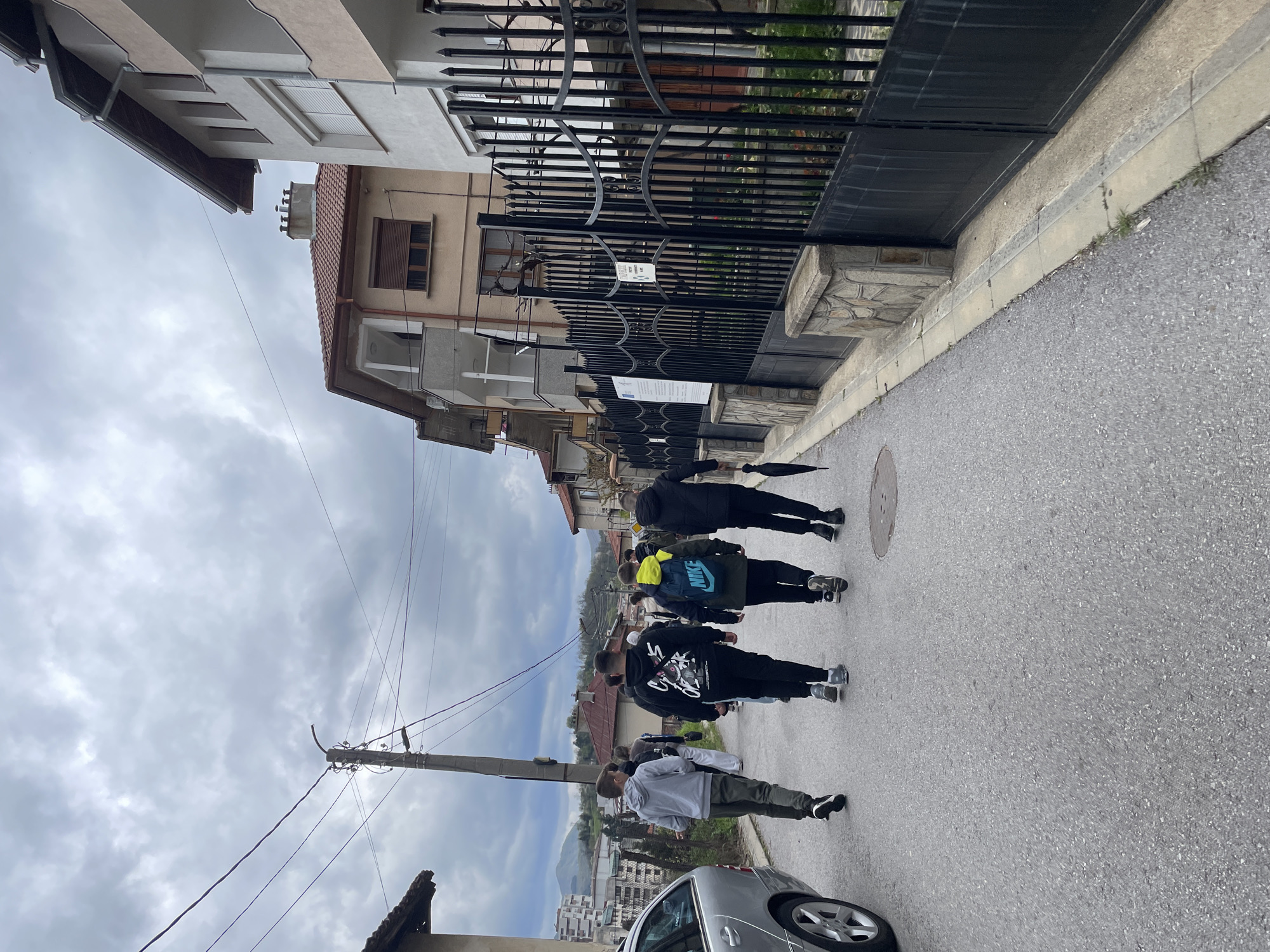 The image captures an uphill street scene in Zlatograd, Bulgaria, under an overcast sky. A group of approximately 5-7 young individuals are walking away from the camera, ascending the paved street. The person closest to the camera wears a grey hoodie and olive pants, while others further up include someone with a blue and yellow "NIKE" branded backpack, another in a black hoodie with white graphics, and one holding a black umbrella.
The street is flanked by buildings of varying architectural styles. On the right, a multi-story building with a light-colored facade is partially obscured by a tall, black ornate metal fence and gate. A sign featuring the "EU" logo and project information, with text likely in Bulgarian, is visible on the fence. Further down the street, another building with a tiled roof is visible. On the left side of the street, more buildings are present, and a utility pole with wires stands prominently, with parts of a silver car (possibly a Mercedes) visible in the foreground.
In the background, rolling hills or mountains are visible, indicating the town's geographical setting. The grey, cloudy sky suggests an overcast day, possibly hinting at recent or impending rain given the umbrella. The scene depicts an everyday activity of people walking along a residential street. No specific event or strong emotions are discernible.