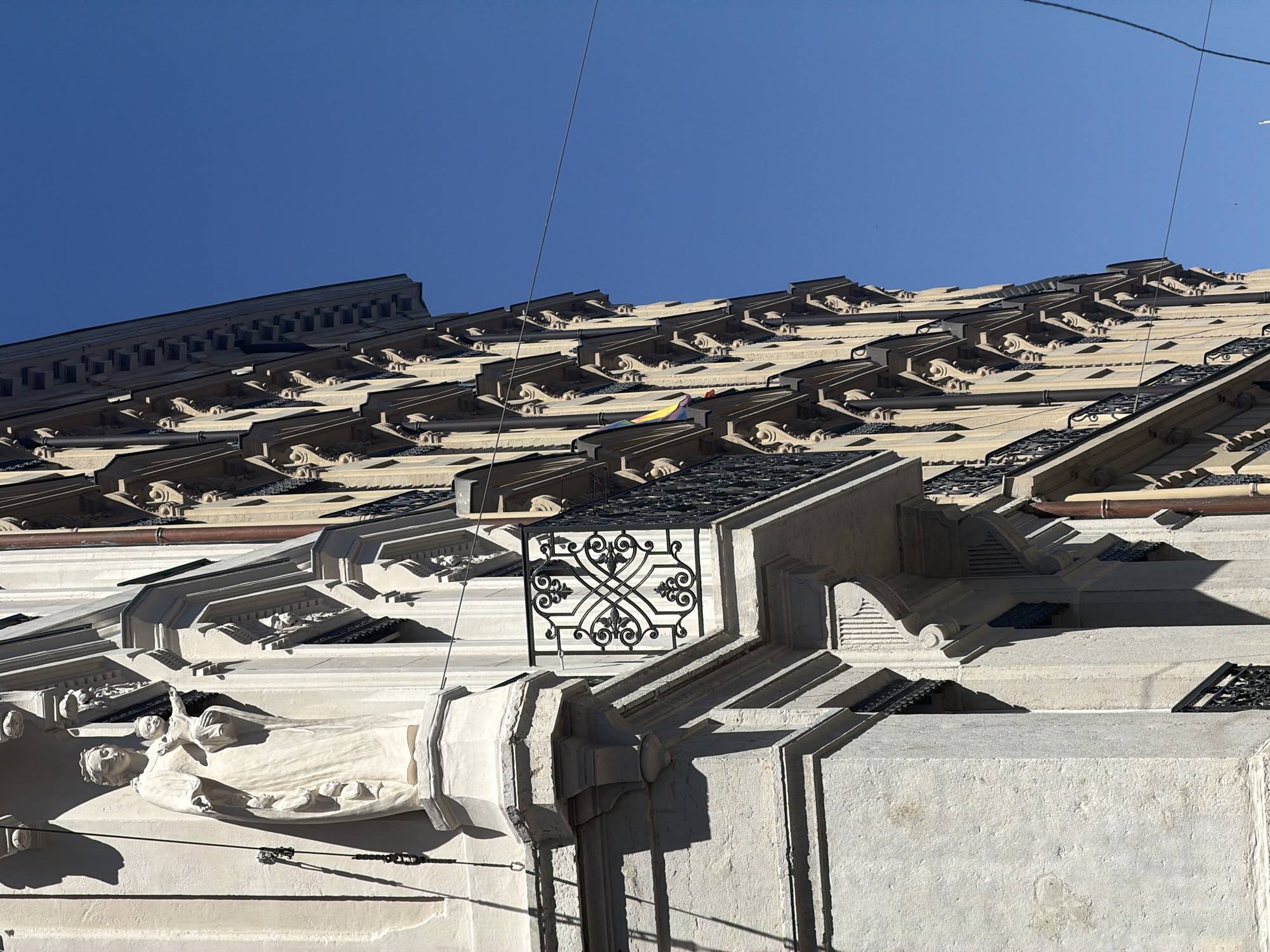 The image shows a low-angle shot of the ornate facade of a building, likely a historic one, with a bright blue sky as the backdrop. The building's architecture features intricate detailing, including decorative carvings, stepped cornices, and metal grilles. In the foreground, a detailed relief sculpture of a reclining figure, possibly a woman holding a child or a mythological representation, is visible. Further up the facade, rows of elaborate decorative elements, resembling stylized acanthus leaves and volutes, are repeated along the roofline. A decorative black metal screen with a swirling pattern is prominently placed on one of the upper levels. The sunlight casts strong shadows, indicating it is daytime, likely mid-morning or afternoon, with clear weather. Some utility wires are visible strung across the sky. The overall impression is one of grandeur and historical significance, suggesting a location in a city with rich architectural heritage. Without more context, the specific activity or event taking place is not discernible, and no visible text is present in the image. Given the provided location context, this building could be in Lyon, France.