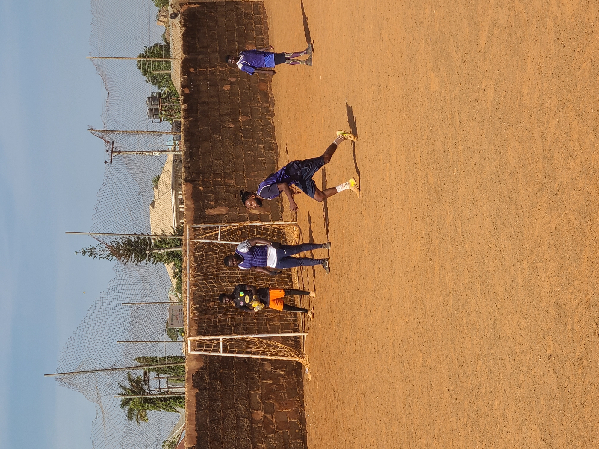 The image shows a soccer field in Barakin Lerawi, Nigeria. Four men are present; two near a goal with netting, one in mid-stride on the field, and another standing in the distance. One individual is wearing a bright orange shirt and gloves, possibly a goalie. The playing surface appears to be dirt. A brick wall runs along one edge of the field, with fencing and vegetation visible in the background. The sky is clear and blue.
