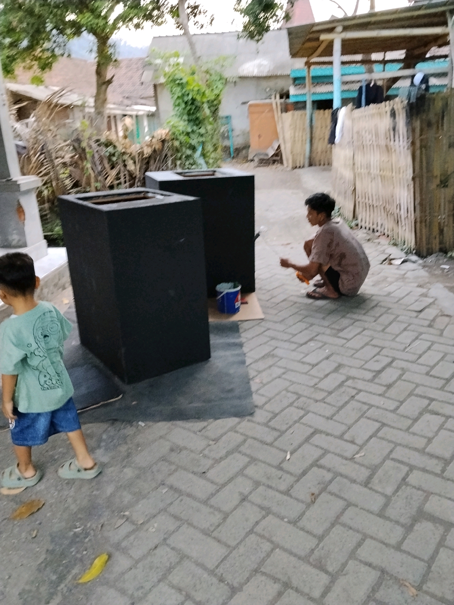 A young boy wearing a mint green t-shirt and blue shorts stands on a paved pathway in Panggungan, Indonesia, next to a tall, black, rectangular object. To his right, a young man squats down, holding an orange object and a paintbrush, appearing to be working on a similar black object. In the background, there are buildings with corrugated metal roofs, bamboo fencing, and trees. The ground is covered with interlocking pavers.