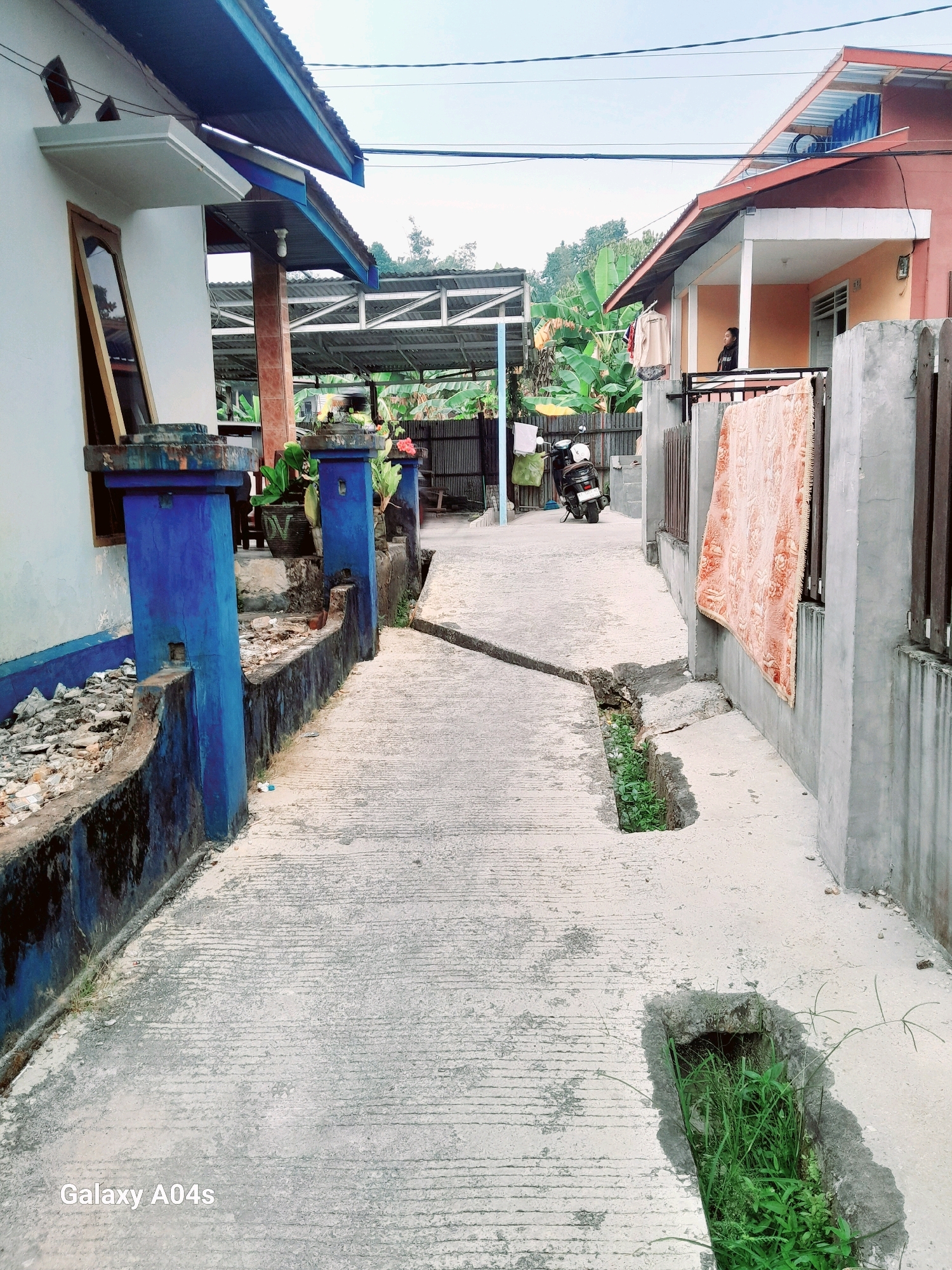 A narrow, paved alleyway runs between two modest houses in Balikpapan, Indonesia. The house on the left is painted white with blue trim, and a dark concrete barrier runs along the side of the alley. The house on the right is painted peach and orange, with a prominent blanket hanging from its exterior wall. A person is visible standing on the porch of the house on the right. A motorcycle is parked further down the alley. Lush green foliage, including banana trees, is visible behind the fence on the right. The sky is overcast. The image appears to be taken with a mobile phone, as indicated by the "Galaxy A04s" watermark in the bottom left corner.