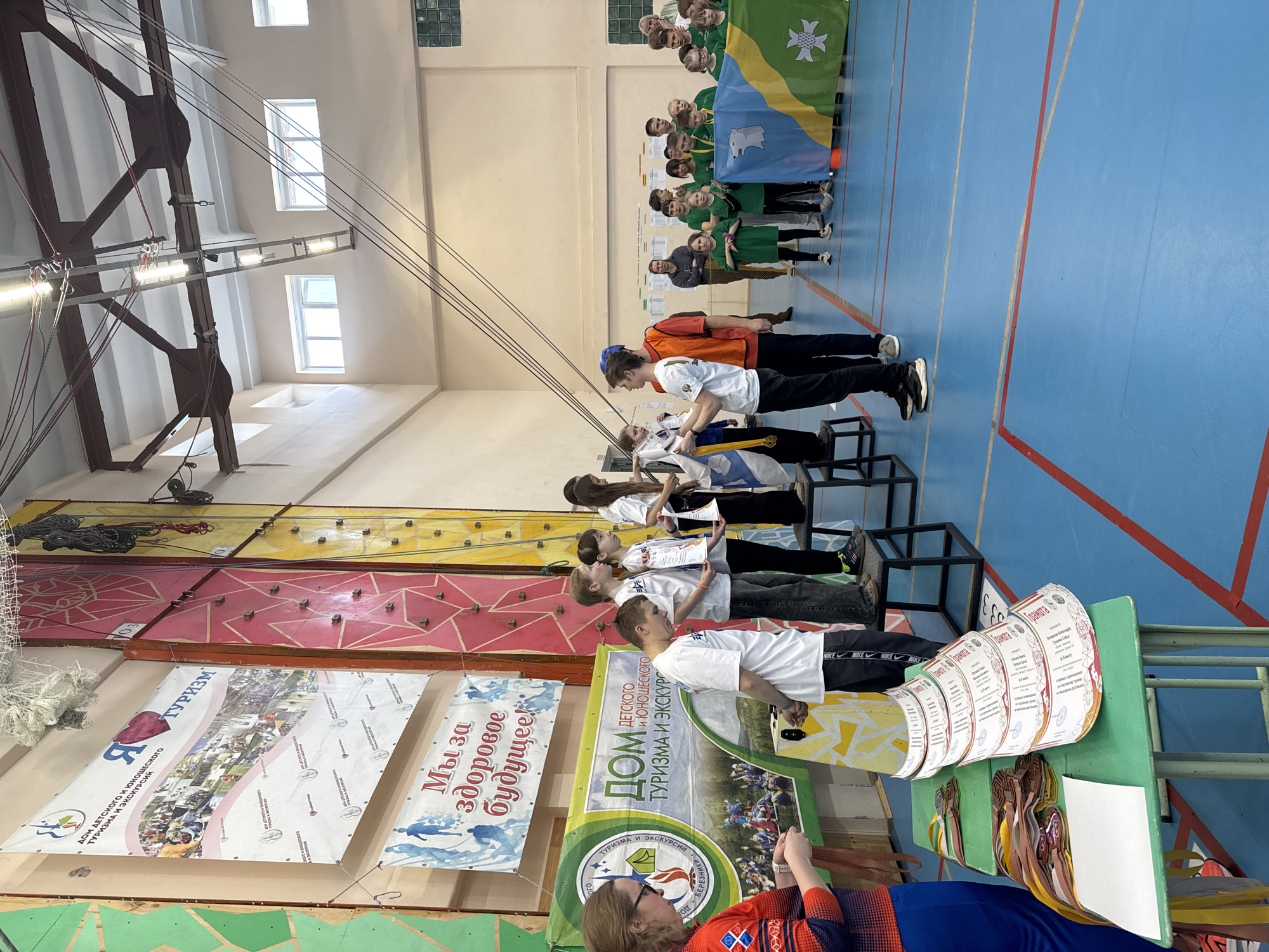 The image depicts an indoor event, likely an award ceremony or celebration, taking place in what appears to be a gymnasium or large hall in Usolye, Russia.
In the foreground, a person with glasses, wearing a red and blue patterned shirt, is taking pictures of a table covered in awards and certificates. The certificates are curled, likely for presentation. A rack holds medals of various colors. Behind the table, a row of children stands on platforms, each holding a certificate, presumably after receiving an award. All the children are dressed in white shirts and dark pants/jeans.
Further back, another group of children is positioned near a wall, holding a flag displaying a green, yellow, and blue design, possibly related to Usolye. The surrounding environment is the hall with blue flooring, a high ceiling, and climbing wall with banners featuring Russian text. Overhead, lighting fixtures are suspended from the ceiling.
