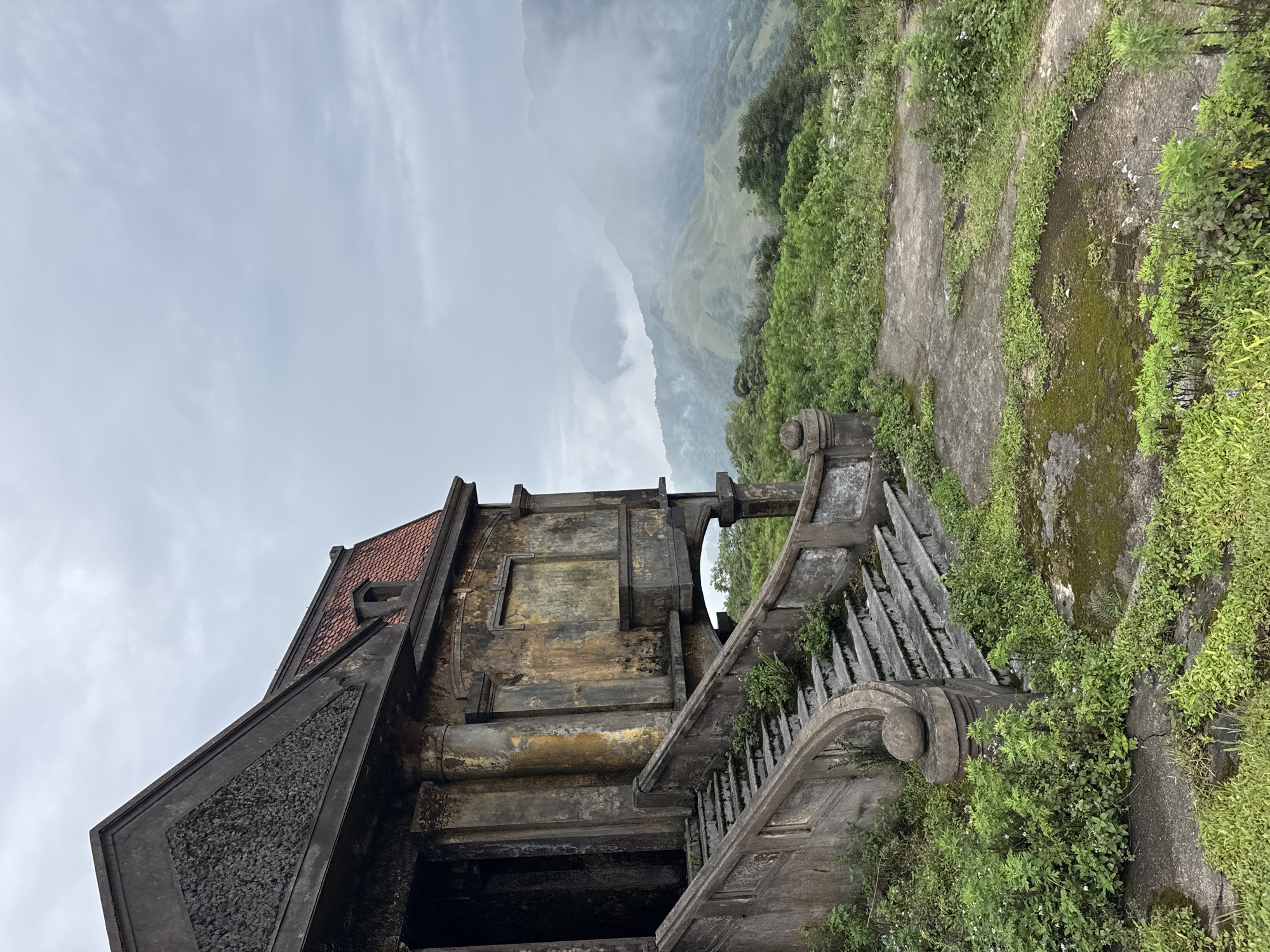 The image depicts a decaying, weathered stone building with a red tile roof, perched precariously on a lush, verdant mountainside overlooking a misty valley. The building's facade is peeling and stained, with a grand, overgrown stone staircase leading up to an arched entrance. The surrounding landscape is characterized by rolling hills covered in dense vegetation, shrouded in a thick, atmospheric fog that obscures the distant mountains. The overall impression is one of abandonment and the slow reclamation of man-made structures by nature in the Mẫu Sơn region of Vietnam.
