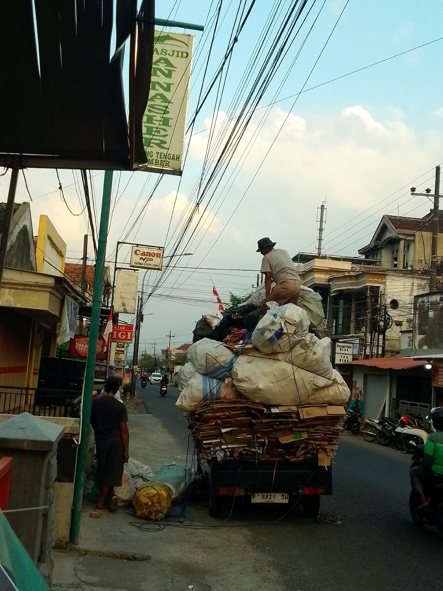 In the city of Gebang, Indonesia, a man wearing a hat and shorts stands on top of a large pile of assorted debris, including large white plastic bags and cardboard boxes, loaded onto the back of a truck. The truck is parked on the side of a paved road, with a sidewalk in the foreground. Scattered on the sidewalk are more plastic bags and debris. A dark-colored utility pole with numerous wires stretches across the frame, obscuring parts of the sky and buildings. Several buildings line the street, some with shops and signs visible, including a sign for "Canon NP TERMA FOTO COPY." Motorcycles are present on the road, and another person stands on the sidewalk near the truck. The sky is partly cloudy.