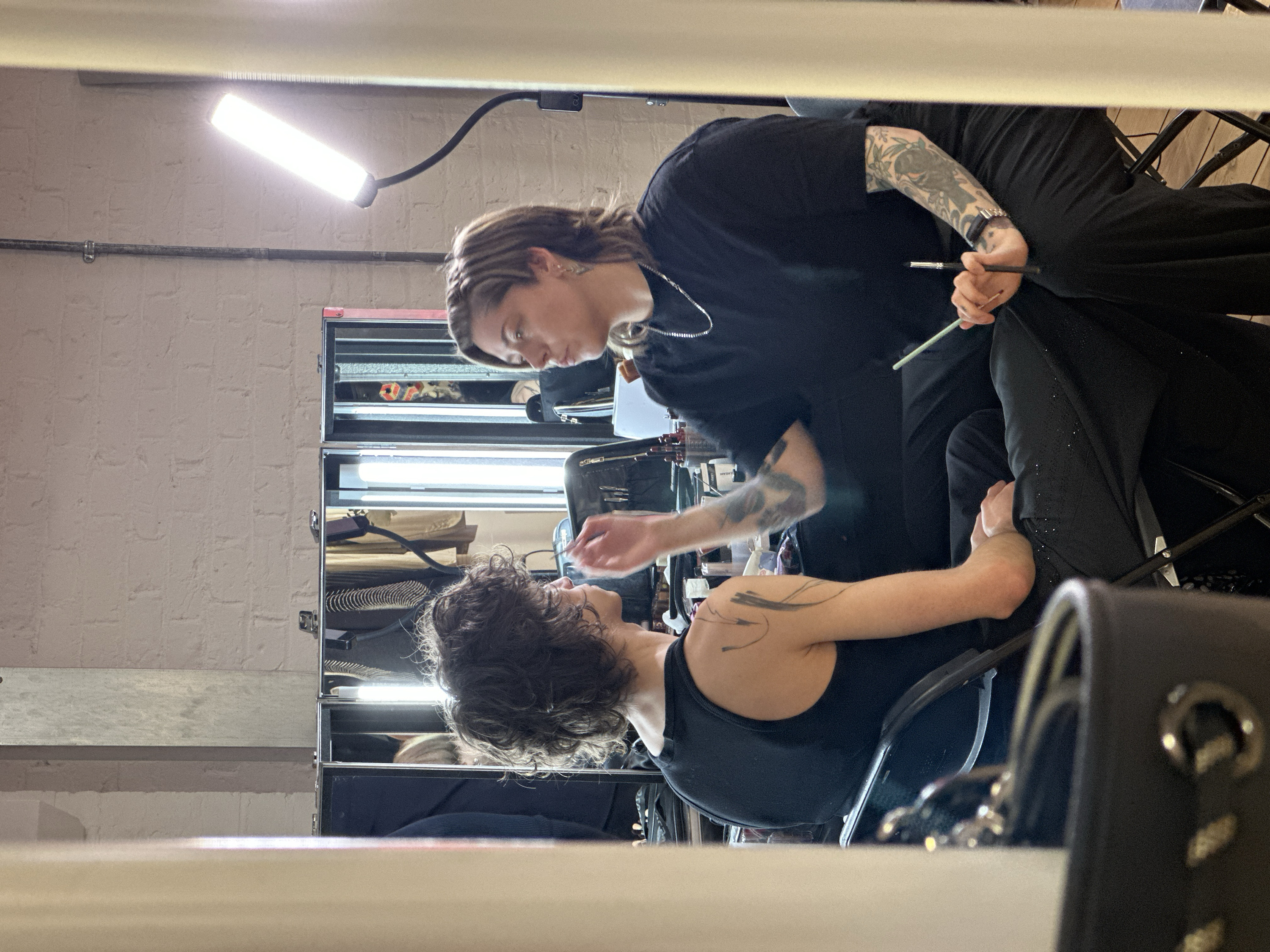 An indoor scene in what appears to be a professional studio or backstage area, located in London, United Kingdom, depicts two individuals engaged in a makeup or body art application.
The subject on the left, with dark, curly hair, is seated in profile, facing right. This individual wears a black tank top, and their visible left shoulder and upper arm display multiple dark, abstract, brushstroke-like markings. Their face is partially visible, oriented towards the subject on the right.
The subject on the right, possessing light brown, layered hair, is also seated, facing left. They wear a black short-sleeved t-shirt and dark trousers, with noticeable tattoos covering both forearms. A silver chain necklace is visible. This individual is actively applying a substance to the left subject's face using a small brush held in their right hand. A second brush is held in their left hand, resting on their lap.
The background features a light-colored, textured brick wall. A metal pipe runs vertically along the left side of the wall. Directly behind the subjects, multiple reflective mirror panels are visible, reflecting various items of clothing, including jackets and striped garments, hung on what appear to be racks or hangers. A tall, flexible LED light panel on a stand provides illumination from behind the right subject. Between the two individuals, an assortment of makeup products, brushes, and containers, including a rack of lipstick tubes, is arranged on a surface, likely a portable makeup station. Both subjects are seated on black folding chairs. The floor appears to be made of wood. The foreground on both the left and right sides of the image is partially obscured by blurry, light-colored vertical elements.
