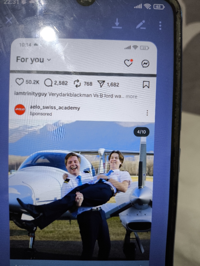 The image displays a dark-colored smartphone screen, oriented vertically, showing an active Instagram feed. The screen exhibits multiple scratches and a significant circular light reflection in the upper left quadrant. The phone's status bar indicates the time as 22:31, full cellular signal strength, Wi-Fi connectivity, and a 63% battery charge.
The Instagram content is a 'For you' section post, labeled as "Sponsored" by "aelo_swiss_academy." The post displays an image, marked as "4/10" in a carousel, featuring two adult Caucasian males positioned outdoors in front of a white single-engine propeller aircraft, likely a Diamond DA40 model. Both men wear pilot uniforms consisting of white short-sleeved shirts, blue ties, and dark trousers; the man on the left displays pilot epaulets. The man on the left, with lighter hair, is holding the man on the right, with darker hair, in a bridal carry position. Both individuals are smiling broadly and laughing. The aircraft's cockpit canopy, propeller, and landing gear are visible. The background consists of a paved surface, green ground, and distant mountains under a clear blue sky.
Below the image on Instagram, interaction metrics show 50.2K likes, 2,582 comments, 768 shares, and 1,682 saves. The post is attributed to "iamtrinityguy" with a partial caption reading "Verydarkblackman Vs B lord wa...".
The image provides no discernible visual information to ascertain its location as Nicosia, Northern Cyprus / Cyprus.