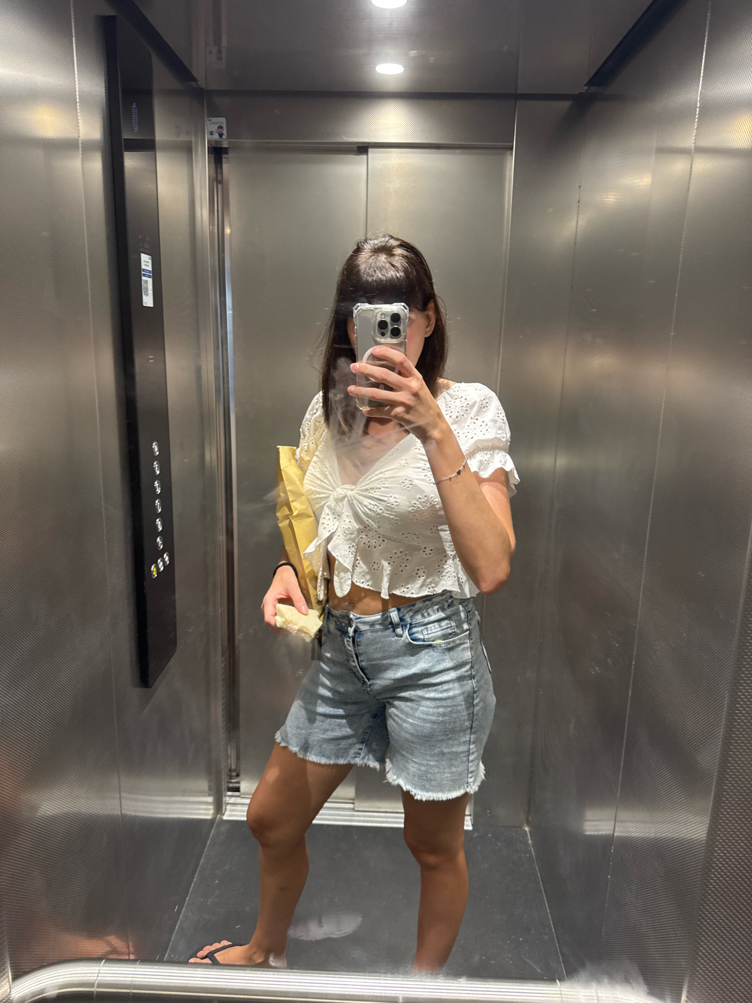 A woman stands in an elevator, taking a mirror selfie. She is wearing a white eyelet crop top with a tie front and ruffled sleeves, and light-wash denim shorts with a frayed hem. She holds a yellow food bag in her left hand and a sandwich in her right. Her hair is dark brown and styled with bangs. The elevator has metallic walls and a control panel with buttons on the left. The floor appears to be dark and reflective. The scene takes place in Isola delle Femmine, Italy.