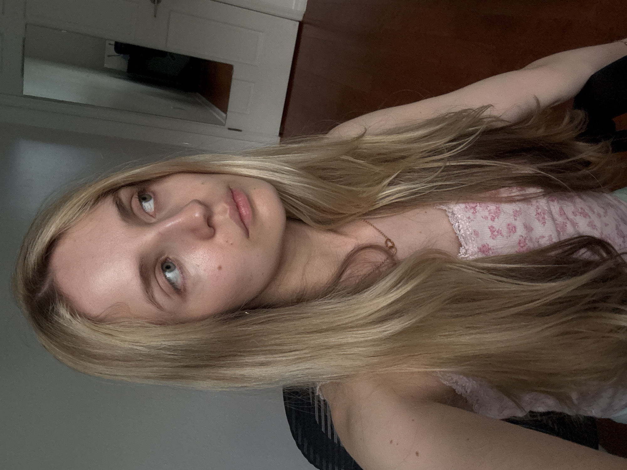 A young woman with long blonde hair is seated and looking off to the right of the frame. She is wearing a pink camisole with a floral pattern. The background includes a gray wall on the left and a doorway with a mirror reflecting a hallway on the right. The floor appears to be dark wood. The image is a close-up of the woman's head and upper body.