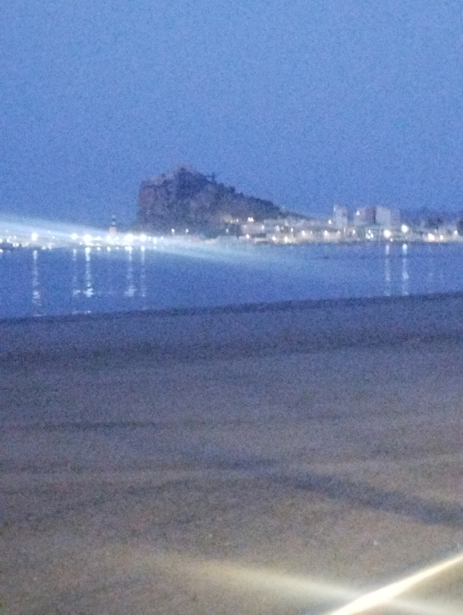 The image depicts a dusky coastal scene with a town lit up in the distance. A prominent rocky promontory, likely Isla de las Larrocas, juts out into the sea, overlooking the town and its lights. The town is characterized by buildings and illuminated structures, suggesting a populated area. The sea reflects the lights, creating a shimmering effect on the water's surface. The foreground is a dark, sandy expanse, possibly a beach, with faint streaks of light suggesting artificial illumination or reflections. The overall atmosphere is serene and tranquil, indicative of twilight or early evening.
