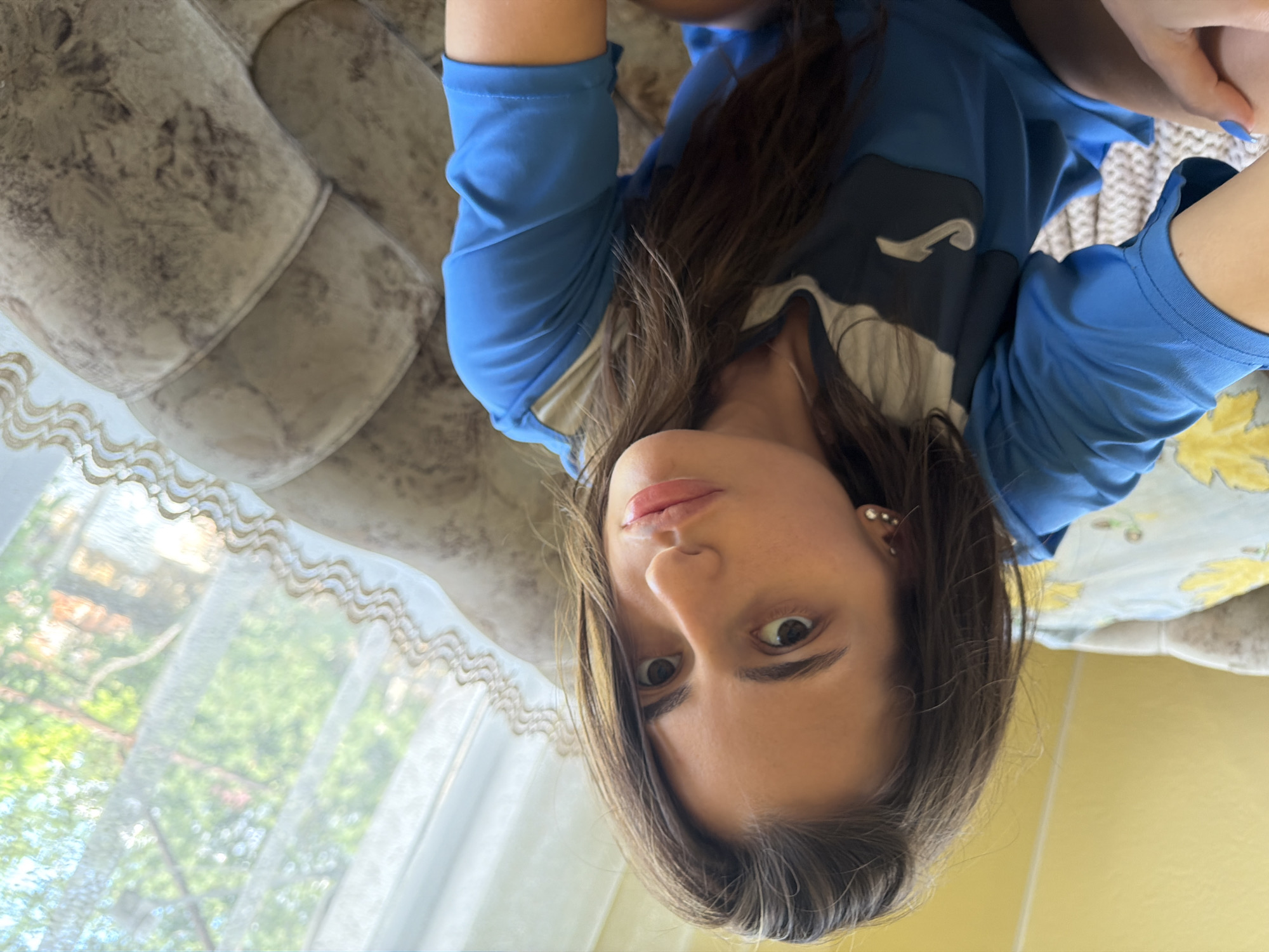 The media file is an upside-down, close-up selfie of a young woman. She is lying on a couch, indicated by the textured upholstery visible in the background. The woman has long, dark hair that falls around her face. She is wearing a blue t-shirt with a dark logo on the chest. Her gaze is directed towards the camera, and her expression appears neutral or slightly curious.
In the background, through a sheer, decorative curtain, greenery and sunlight suggest an outdoor scene. The lighting indicates it is daytime, likely bright. The overall setting appears to be a living room or a comfortable indoor space. The image is oriented such that the woman's head is at the bottom and her feet would be at the top. There is no visible text in the image.