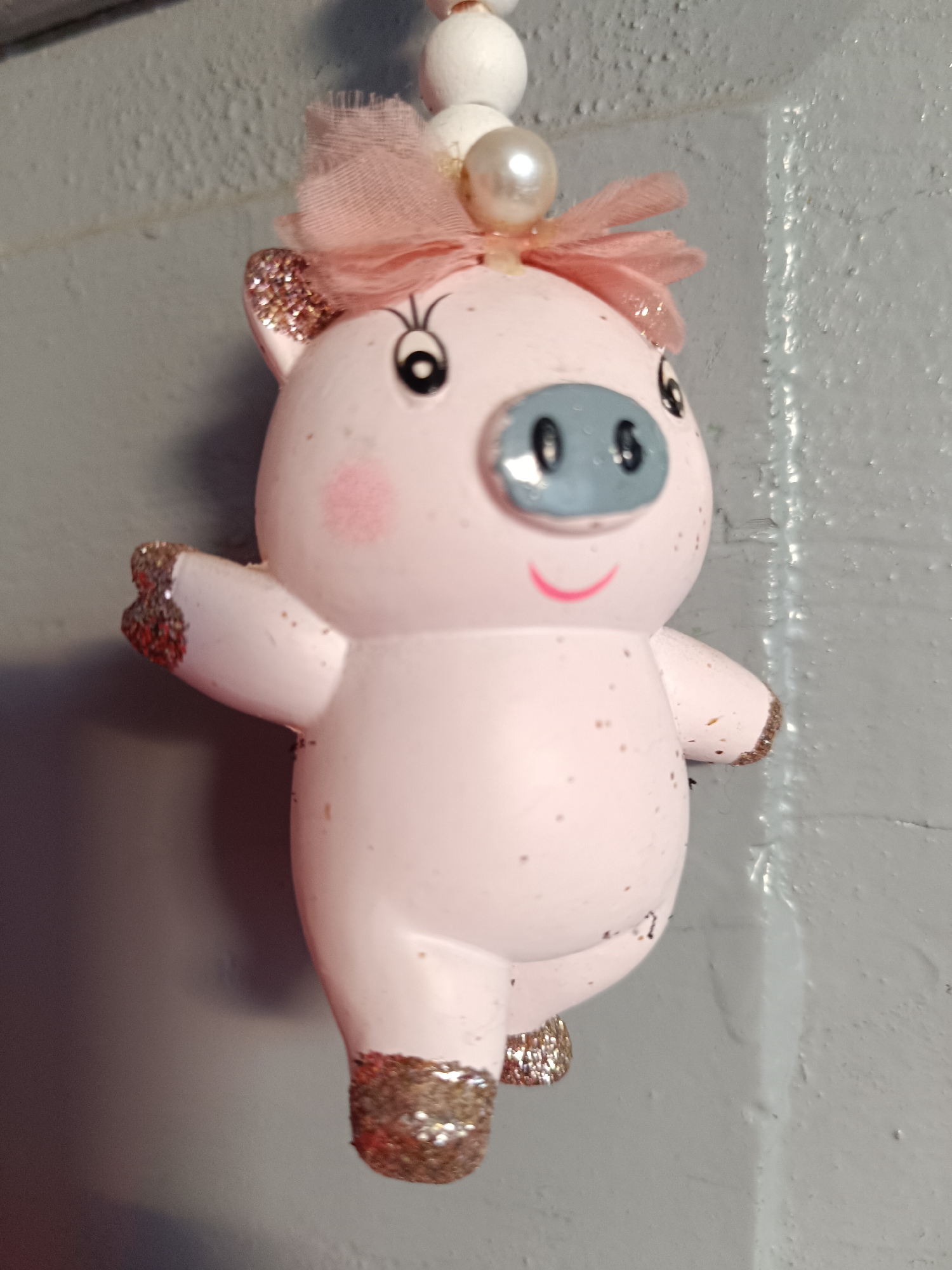 A light pink, glitter-dusted ceramic pig figurine is suspended against a gray, textured wall. The pig has a blue snout with black nostrils, black dot eyes, and a pink smile. A pink bow with a pearl embellishment adorns its head. The figurine is decorated with gold glitter on its ears, hands, and feet. The background appears to be an indoor wall, likely in a residential setting. No specific location in Tebras, Latvia can be determined from the image.