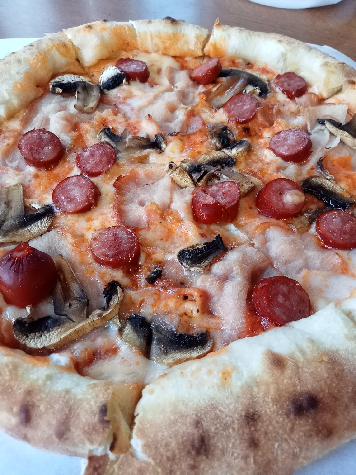A close-up, overhead shot showcases a delicious-looking pizza. The pizza has a golden-brown, slightly puffy crust, topped with melted cheese, slices of red sausage, and pieces of mushroom. Some of the sausage slices are slightly charred at the edges, and the cheese appears melted and gooey. The background consists of a light-colored surface, possibly a table. The overall impression is of a freshly prepared and appetizing pizza ready to be enjoyed.