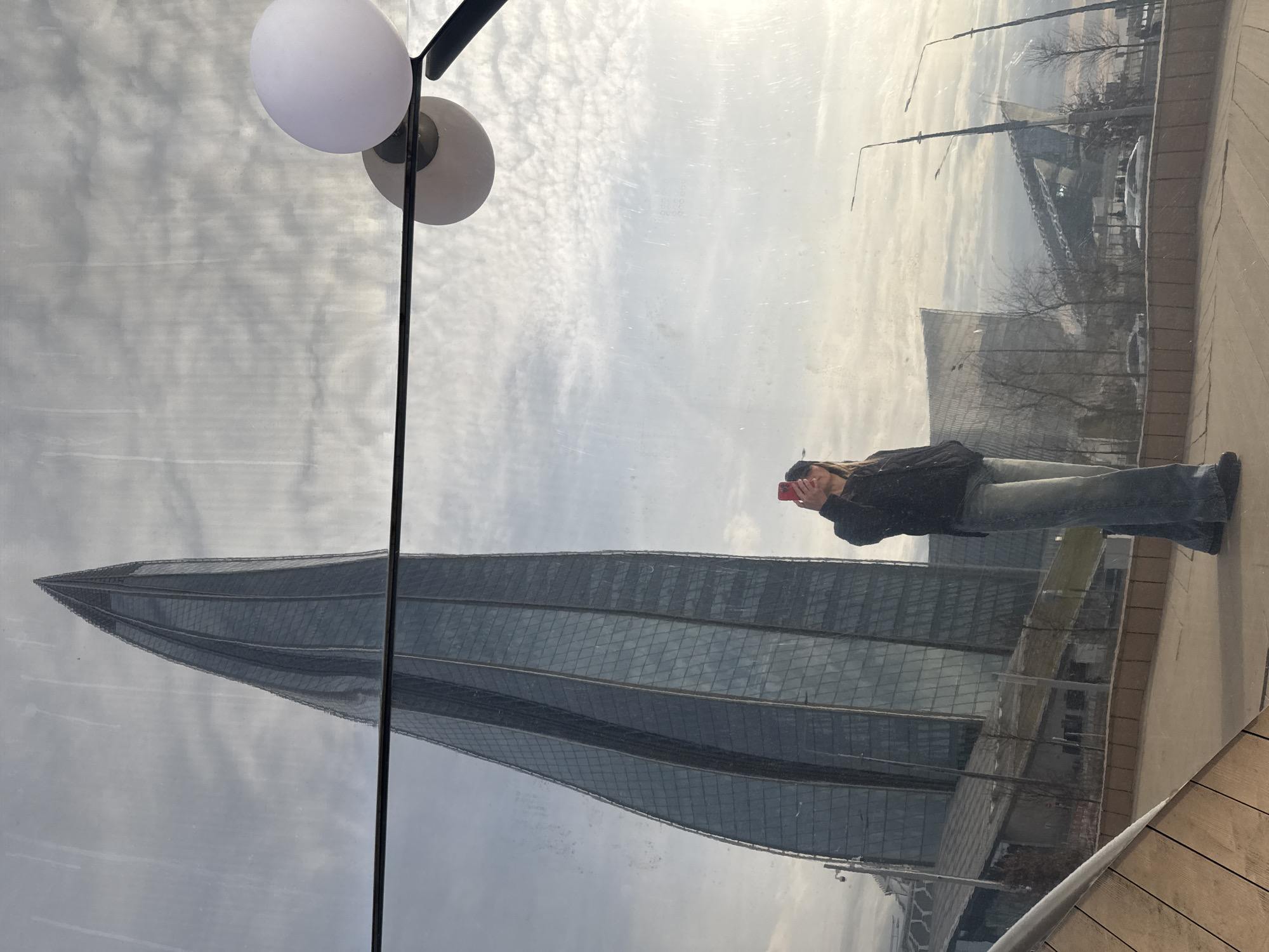 The image is a reflection in a window, likely taken indoors. The main subject of the reflection is the Lakhta Center, a skyscraper in Saint Petersburg, Russia, which is identifiable by its unique pointed shape and glass façade. In the reflection, there is also a bridge visible in the background, possibly one of the bridges over the Neva River. A person is also reflected, holding a red phone as if taking a picture of the building. A lamp with two round, white lights is visible in the foreground, appearing as part of the indoor setting from where the photo was taken. The surrounding environment appears to be an overcast day, and the surface where the person is standing is a wooden deck or walkway.