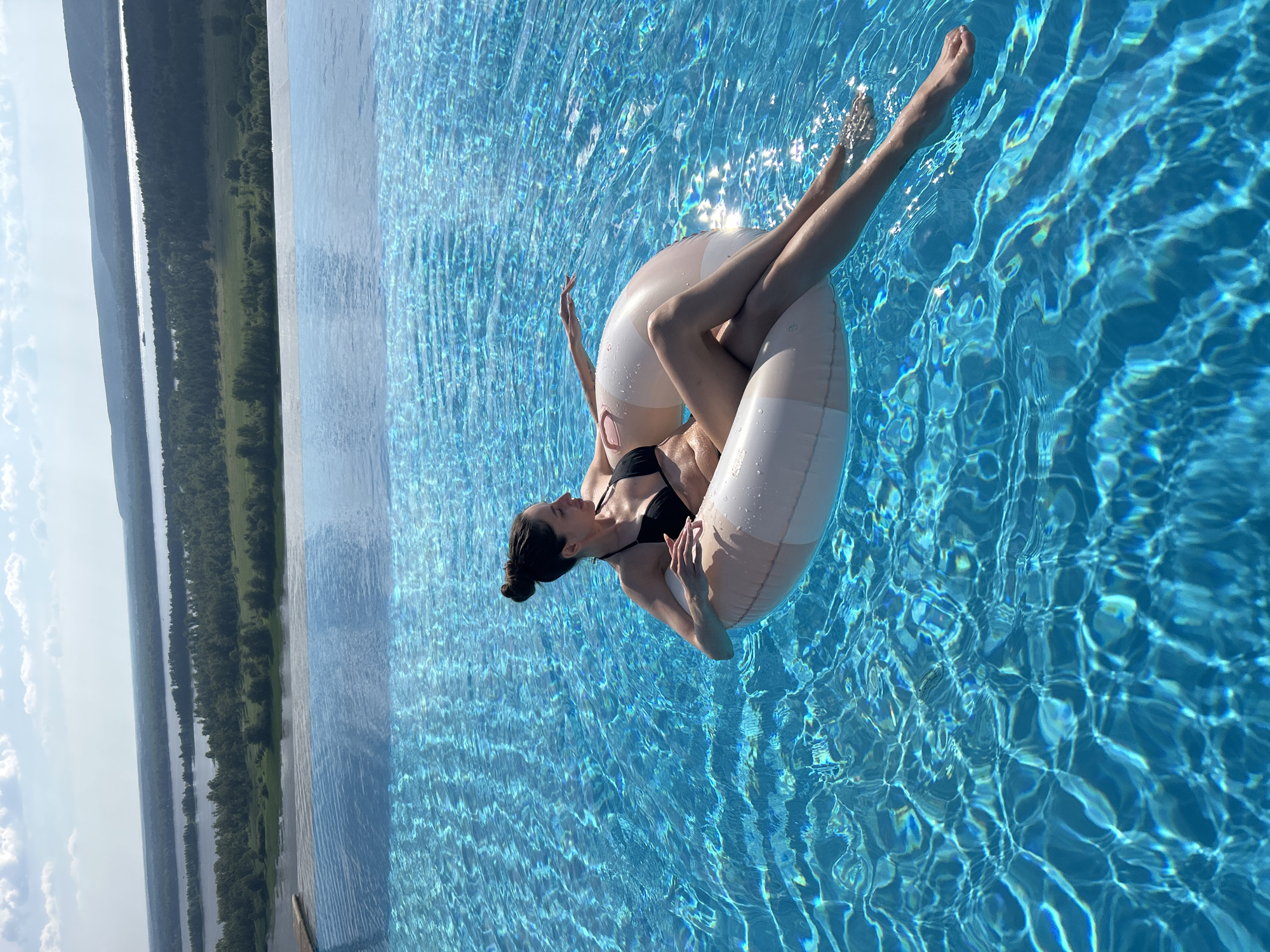 A person is floating on an inflatable tube in a swimming pool. The person is wearing a black bikini and has their hair pulled back in a bun. They are lying on their back, with their legs extended and their arms resting on the tube. The pool is filled with clear blue water, and sunlight is reflecting off the surface. In the background, there is a landscape with a body of water and trees. The image is taken from a high angle, looking down into the pool. The scene appears to be set on a sunny day, likely during the daytime. The overall mood of the image is relaxed and peaceful. The location appears to be a resort or a private residence with a scenic view.