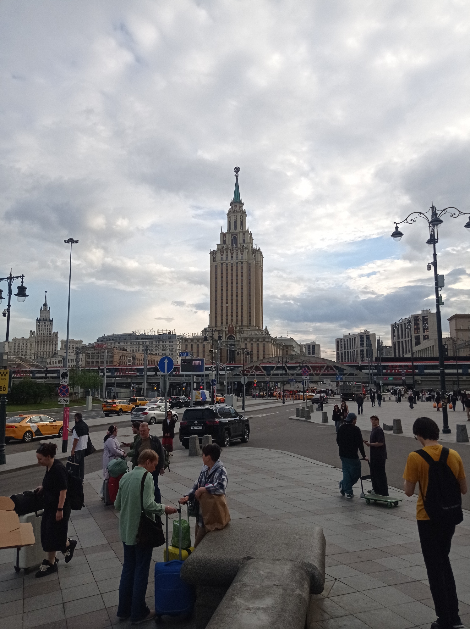 This is a wide-angle, eye-level, full-shot photograph of a street scene in Moscow, Russia, under a cloudy sky. The iconic Stalinist skyscraper, the Ministry of Foreign Affairs building, dominates the background, with its distinctive spire topped with a star. In front of it are other buildings, a raised railway track with a train, and various signs and traffic signals.
In the foreground, a paved walkway is populated by several people with luggage. Some are standing or walking, while others appear to be waiting. To the left, a woman in a black dress is standing next to a cart with boxes and a large black bag. In the middle ground, a woman in a green top and blue pants is standing with a suitcase and a green bag on wheels, while another woman in a plaid jacket stands next to her. To the right, a young man in a yellow shirt and black backpack is walking away from the camera. Further back, several individuals are interacting, including two men, one of whom is pushing a cart with a skateboard.
The street itself is busy with traffic, including several yellow taxis and other cars. Some vehicles are stopped at intersections or traffic lights, suggesting a degree of congestion. The overall impression is of a bustling urban environment, likely a transportation hub, given the presence of travelers and luggage. The weather appears overcast, with visible clouds in the sky.