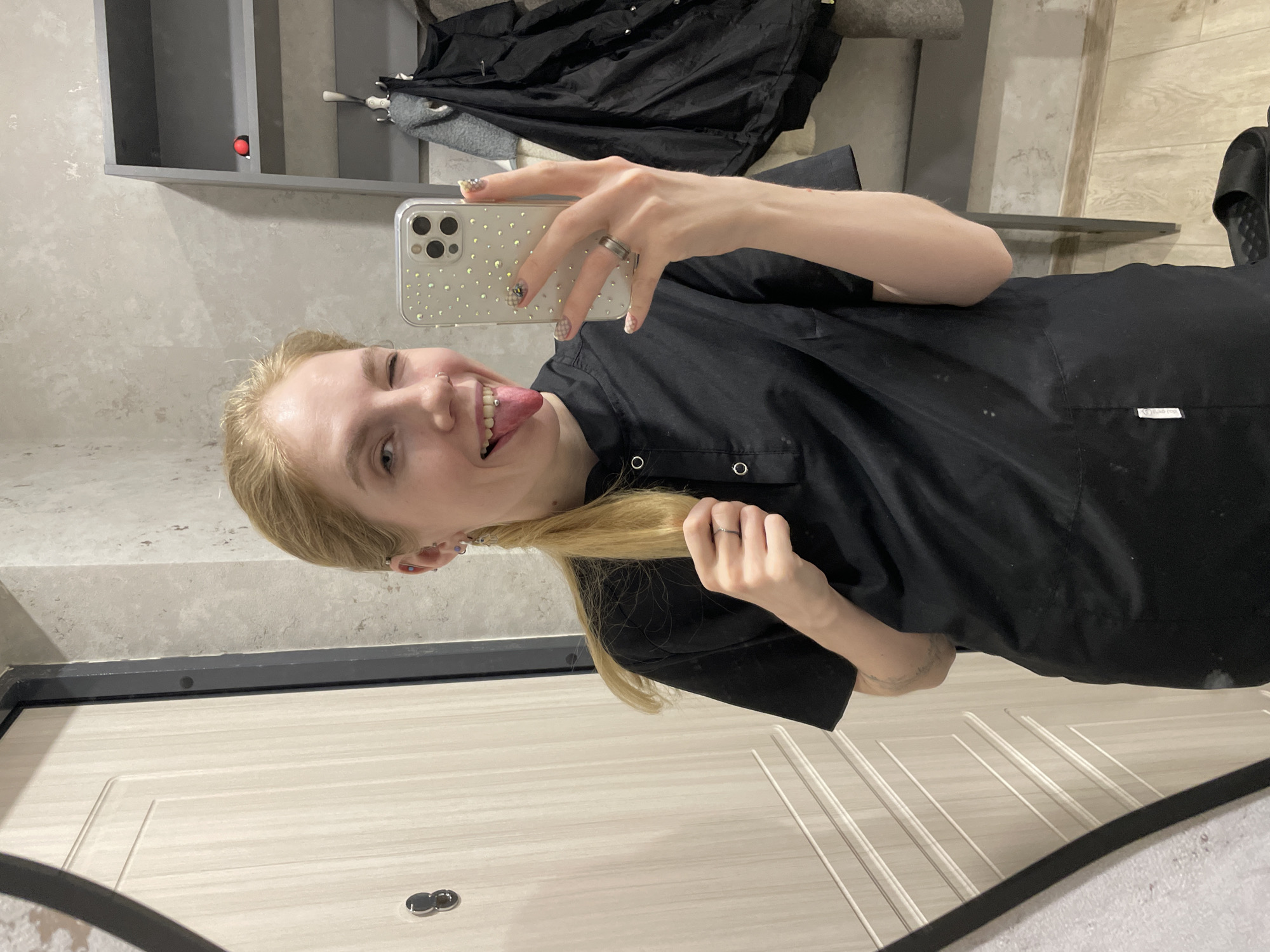 A young, fair-skinned person with blonde hair, wearing a black button-down shirt, is taking a selfie in what appears to be an entryway or hallway. They are sticking their tongue out and holding a section of their hair. The person is using a smartphone with a bedazzled case. The background shows a light-colored door, a gray wall, and a shelf with clothes hanging on it. It is not possible to determine the specific location in Izhevsk, Russia from the given information.