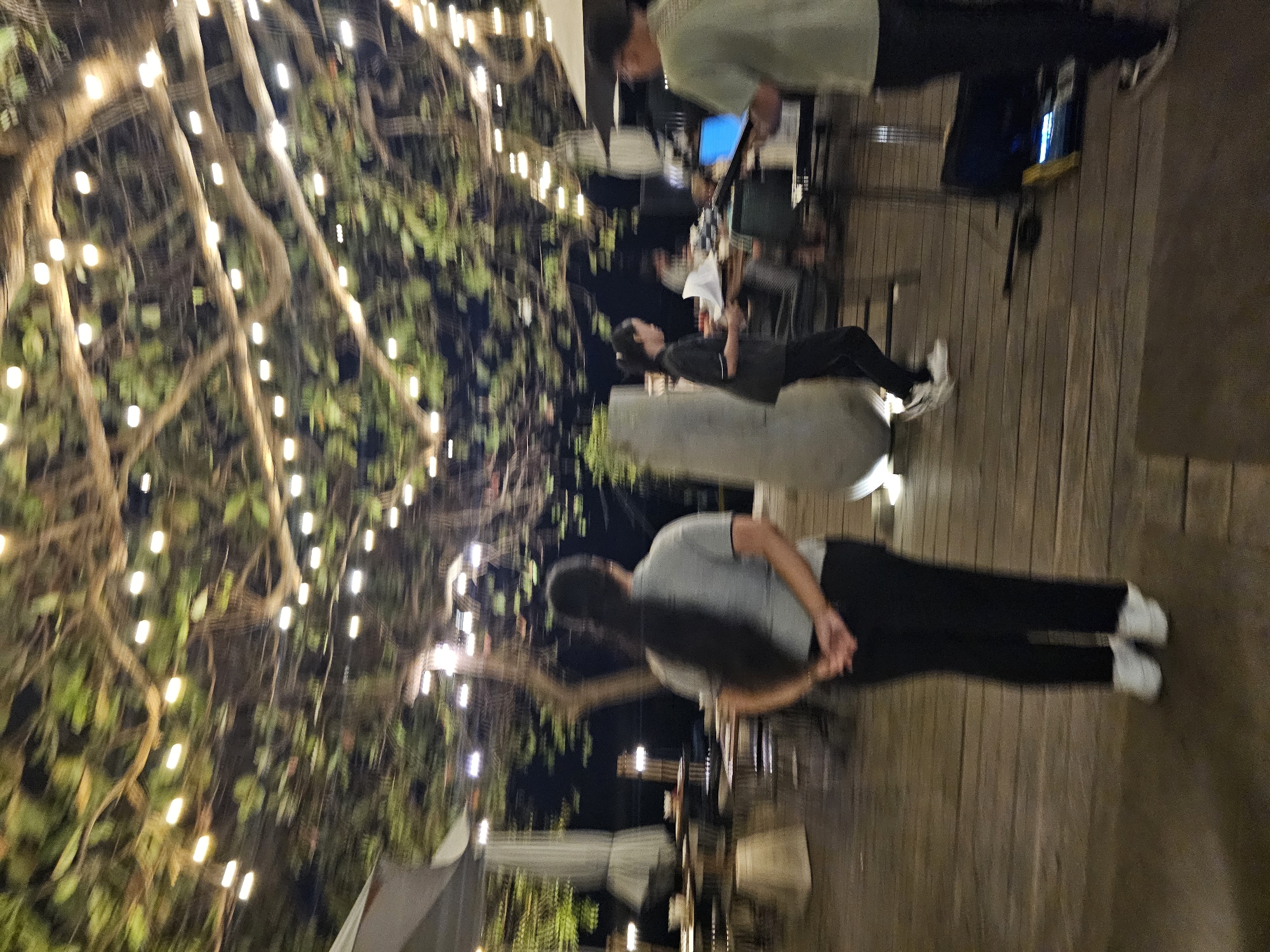 This is a nighttime, slightly motion-blurred photograph taken at an outdoor venue in Denpasar, Indonesia, likely a restaurant or event space, characterized by a wooden deck under a canopy of trees strung with decorative lights. In the foreground, a woman with long dark hair, wearing a grey polo shirt and black pants, stands with her hands clasped behind her back, facing away from the camera. To her right, a younger woman in a dark uniform and black pants is walking briskly, carrying some items, possibly a server or staff member. Further in the background, to the right, a man wearing a light green t-shirt and dark pants is visible, appearing to interact with electronic equipment, possibly a DJ setup or sound system, indicated by a laptop and speakers. The environment features outdoor seating with tables and chairs partially visible, along with large white umbrellas that suggest shelter. The overall ambiance is relaxed and social, illuminated by the warm glow of numerous string lights woven through the branches overhead, creating a festive atmosphere.