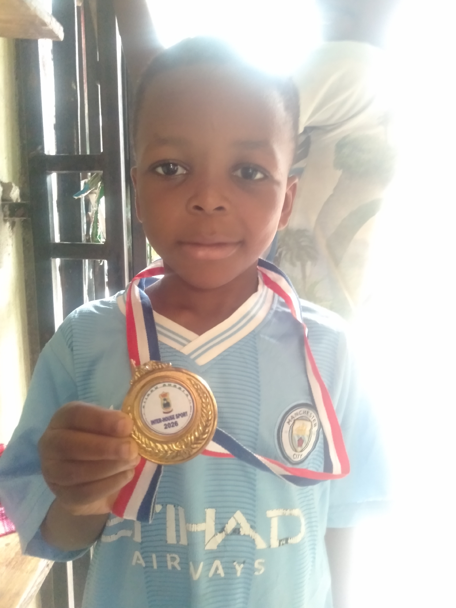 A young boy, likely of primary school age, is captured in a close-up portrait, proudly displaying a gold medal. He is wearing a light blue Manchester City football club jersey with white striped details on the collar and the club's crest on his left chest. The "ETIHAD AIRWAYS" sponsor logo is also visible across the front of the shirt. The medal hangs around his neck with a red, white, and blue striped ribbon, and he holds the medal slightly forward with his right hand. The circular insert on the gold medal clearly reads "INTER-HOUSE SPORT 2026" along with a small crest.
The boy looks directly at the camera with a gentle, proud smile, indicating happiness with his achievement. The setting appears to be an indoor or semi-enclosed space, possibly a doorway or passage, in Shangisha, Nigeria. To the left and behind him, a dark metal security gate or grille is visible against a light-colored wall. The image is brightly lit, with strong light source from the background creating some overexposure, particularly on the right side of the frame and around the boy's head. A faint, blurry outline of another person or object is visible in the bright, washed-out background area to the right.