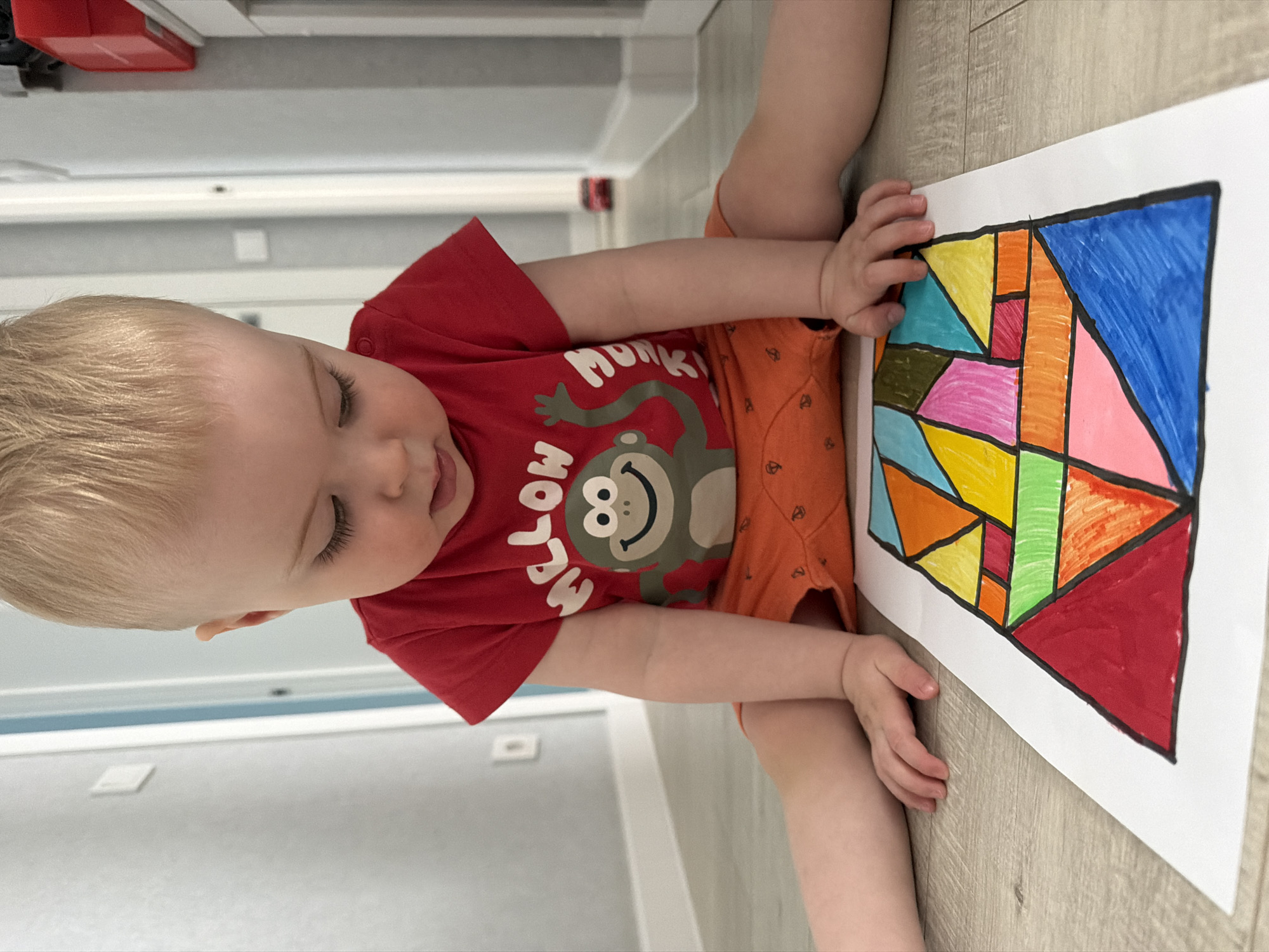 A young child sits on a wood-look floor, presumably indoors given the neutral-toned walls and baseboards visible in the background. The child has fair hair and is wearing a red T-shirt with a monkey graphic and orange shorts. The child is looking at a large piece of white paper on which they have colored a geometric design with various colors including blue, red, orange, yellow, green, and pink, outlined in black. The child appears to be touching the paper.
I have no information whether the photo was taken in Votkinsk, Russia.