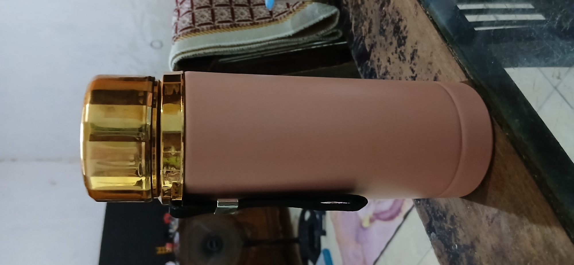 A pink thermos with a gold lid and a black strap is on a wooden table. The background is blurry but shows a fan and a rug. The scene appears to be indoors, possibly in a home in Samarinda, Indonesia.