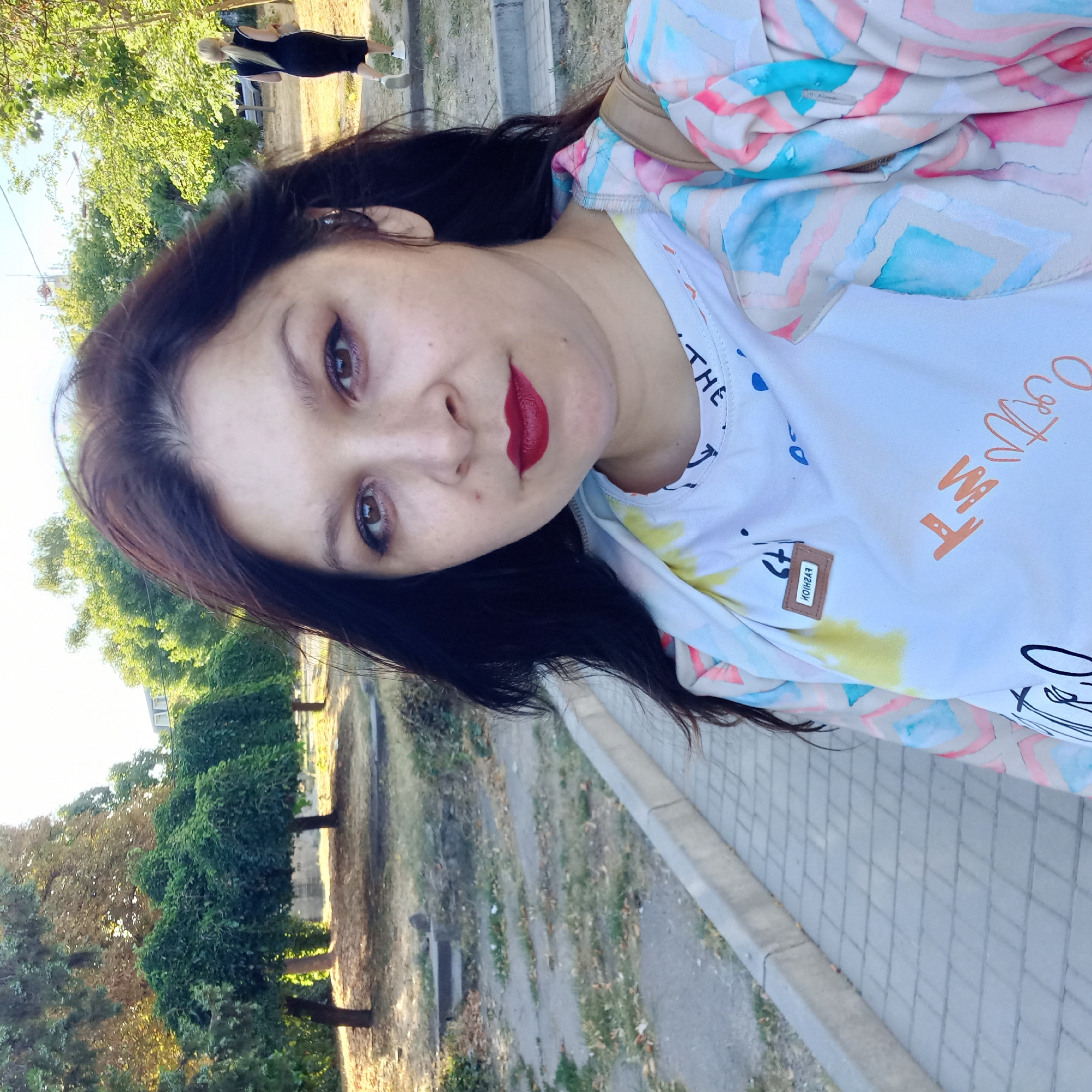 A woman with dark hair and red lipstick is taking a selfie outdoors in Kropyvnytskyi, Ukraine. She is wearing a white t-shirt with colorful graphics and a pastel-colored jacket with a geometric pattern. In the background, there are several trees with rounded tops, a paved walkway with a curb, and a blurred figure of another woman in a black dress walking away. The overall scene suggests a park or a pedestrian area.