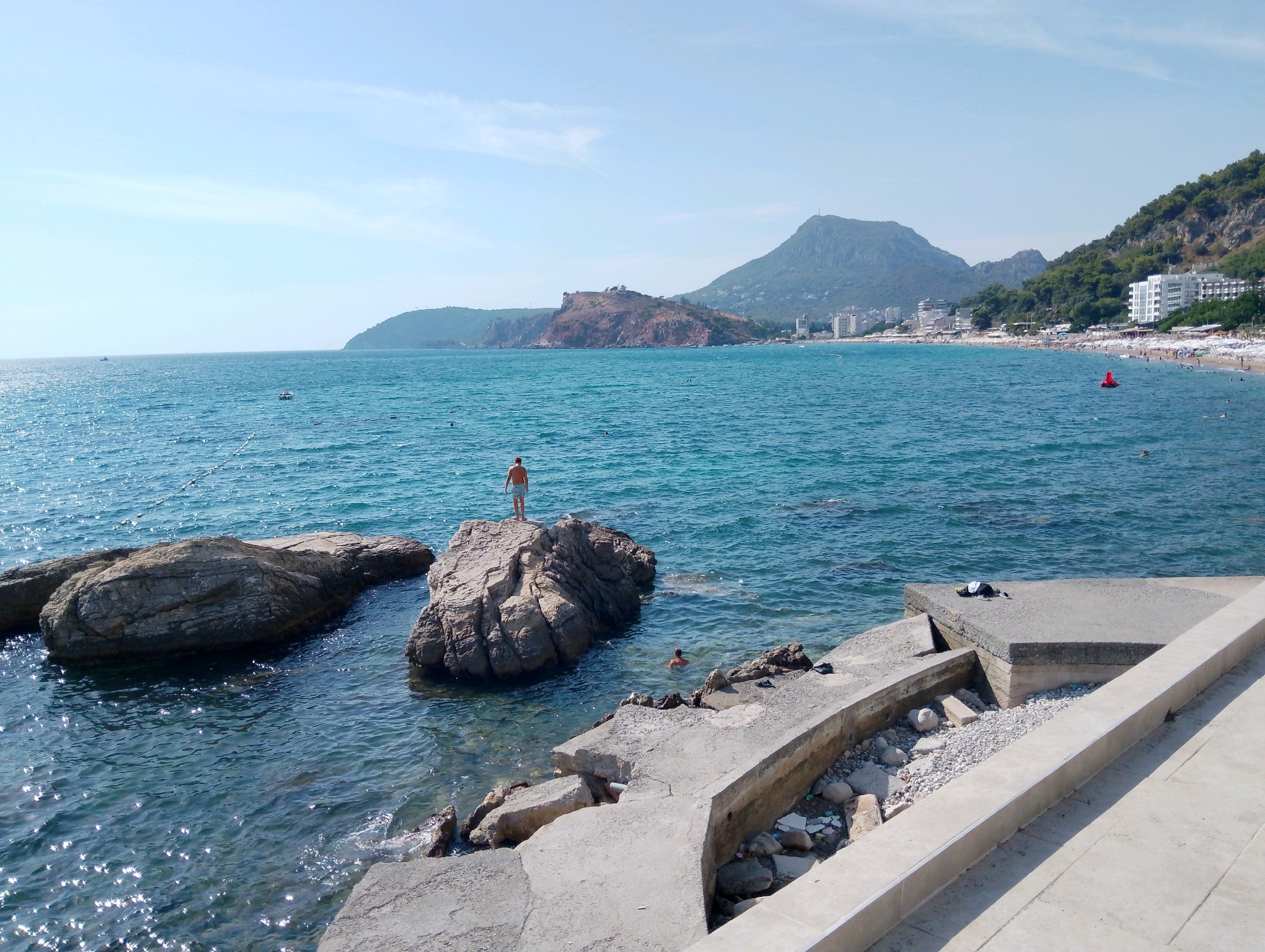 The image shows a coastal scene in Sutomore, Montenegro. In the foreground, a concrete structure with some debris lines the edge of the water. Several large rocks are partially submerged in the clear, blue sea. On one of the rocks, a man stands, facing away from the camera. Another person is in the water near the rocks. In the middle ground, the turquoise sea stretches out, with a few small boats visible. In the background, a beach with numerous umbrellas and sunbathers is visible, backed by a hillside covered in trees and dotted with buildings. Further in the distance, mountains rise, one of them with a settlement on its slopes and what appears to be a communication tower on its peak. The sky is clear and blue with a few wispy clouds. The overall scene depicts a sunny day at a popular seaside resort.