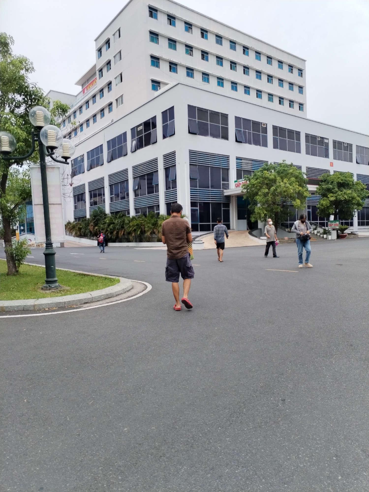 The image shows a paved area outside a white building with multiple floors and many windows. Several people are walking in the paved area. In the foreground, a man with a brown shirt and shorts is walking away from the camera. There is a lamppost on the left side of the frame. The building has signs in Vietnamese. The sky is overcast. The location is in the city of Nha Trang Ward, Vietnam.