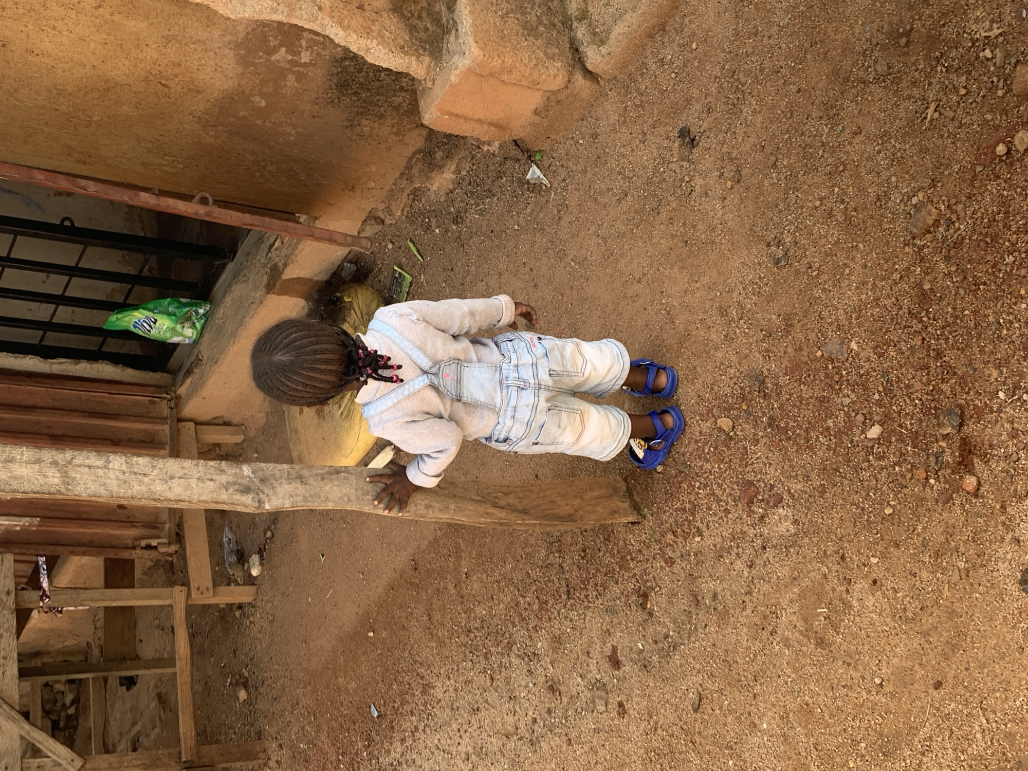 A young child with braided hair stands in a dirt area in front of a building. The child is wearing light blue overalls and a light-colored long-sleeved shirt, with blue sandals. The child's left hand is on a wooden post. In the background, a green bag is visible through a barred window. The ground is a mixture of dirt and small stones. The scene appears to be outdoors, possibly in a residential or market area in Jos, Nigeria.
