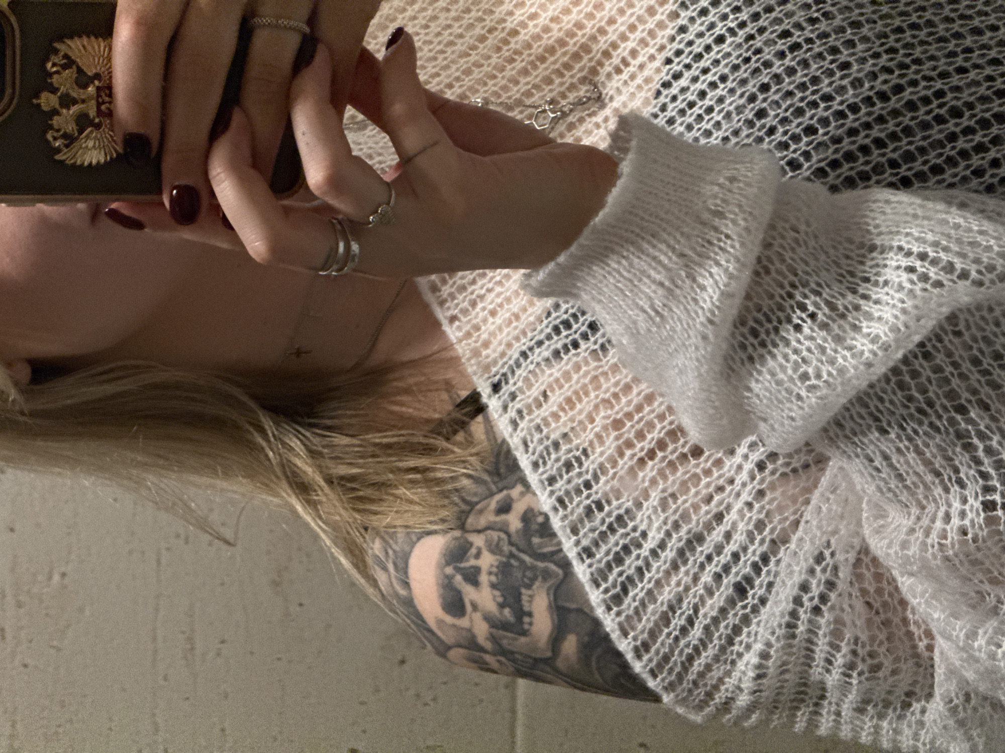 The image shows a person holding a dark-colored smartphone with a gold-colored emblem featuring a double-headed eagle. The person has light blonde hair and a tattoo of skulls on their upper arm. They are wearing a white, loosely knitted garment. The person is also wearing multiple rings on their fingers and a delicate necklace. The background appears to be a plain, light-colored wall. There is no information provided in the image to indicate this scene is from Krasnoyarsk, Russia, or any other location.
