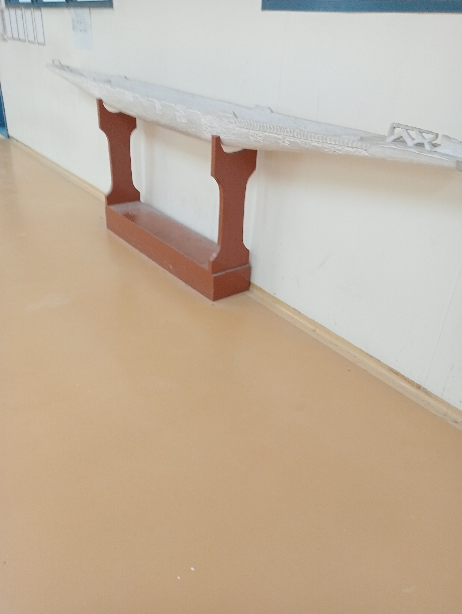 A light-colored, elongated object, possibly a canoe or sculpture, is displayed on a reddish-brown wooden stand against a white wall. The object has intricate patterns carved into its surface. The floor is a smooth, light brown material. The scene appears to be indoors, with the wall showing some minor scuff marks and a baseboard along the floor.