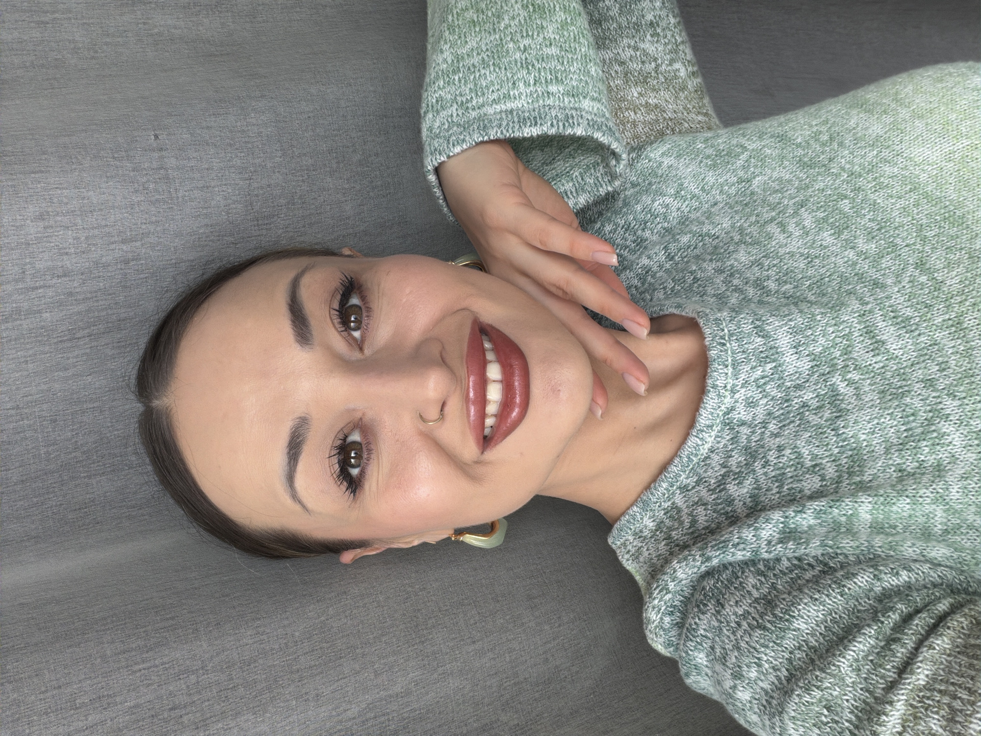 A woman lies on a gray surface, possibly a couch or bed, wearing a light green sweater. She has brown hair pulled back, dark brown eyes, and a nose piercing. Her lips are glossy with dark red lipstick, and she is wearing large gold heart shaped earrings. She smiles and her hand is touching her neck. The location is not provided in the image.