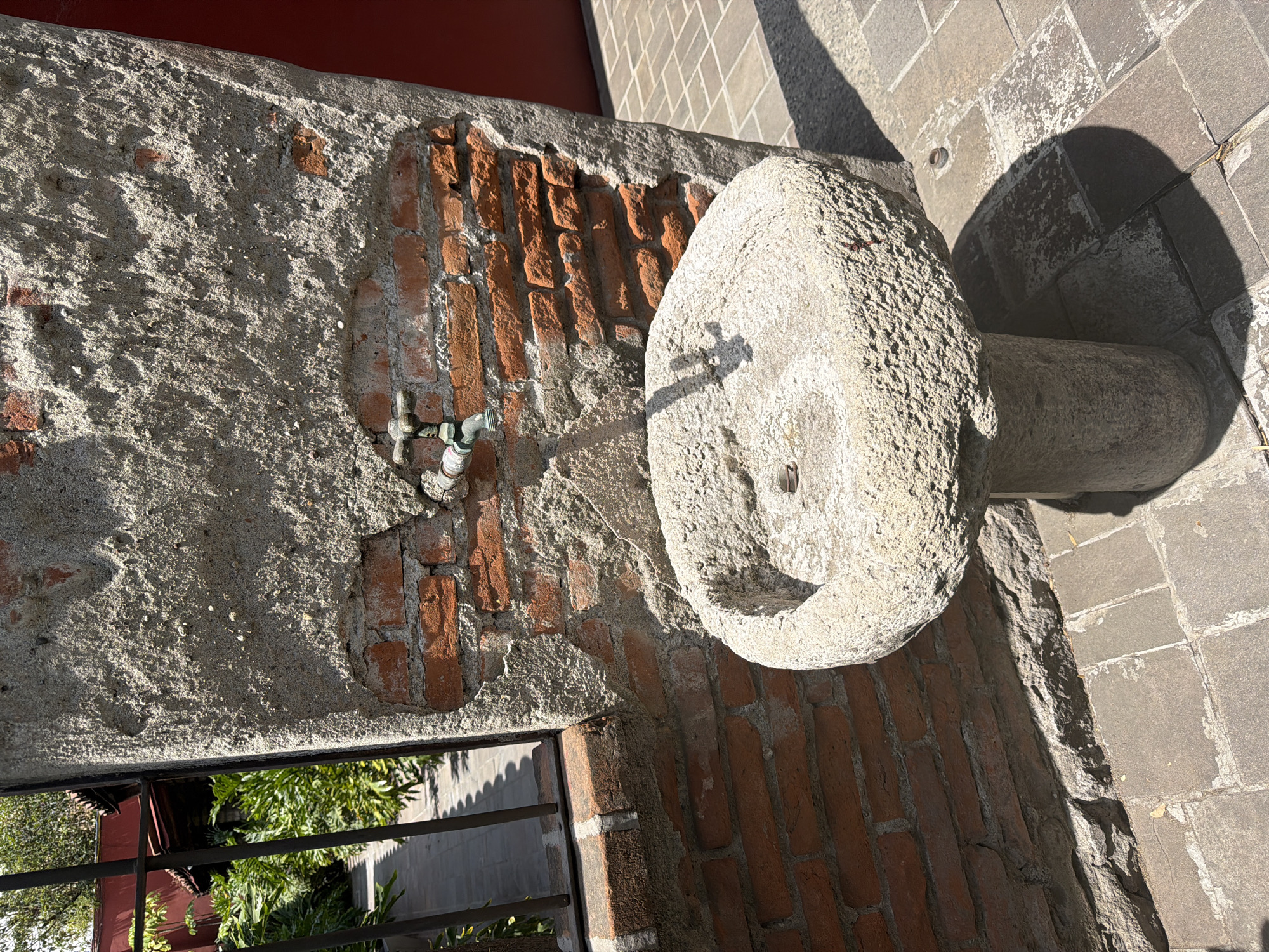 The image displays an outdoor scene, likely a courtyard or passage, under bright, direct sunlight. The perspective is rotated, showing a weathered wall stretching across the left and bottom of the frame, constructed from red bricks partially covered in rough, light grey plaster or cement. An aged metal spigot is affixed to this wall.
To the right of the wall stands a rustic stone fountain, featuring a shallow, circular basin resting on a cylindrical pedestal, both sculpted from textured, light-colored stone. The strong sunlight casts a distinct shadow of the spigot onto the fountain's basin and a large, rounded shadow of the fountain itself onto the ground. The ground is paved with square or rectangular grey stones or tiles.
Above the main stone wall, a smooth, dark red surface is visible, possibly part of another building or structure. At the bottom left, an opening, framed by dark metal bars, reveals lush green foliage and hints of distant structures. The scene is static, devoid of human activity. The harsh shadows and bright illumination suggest a clear, sunny day. The materials and architecture evoke a sense of age and traditional design, fitting the context of Puebla, Mexico. There is no visible text in the image.