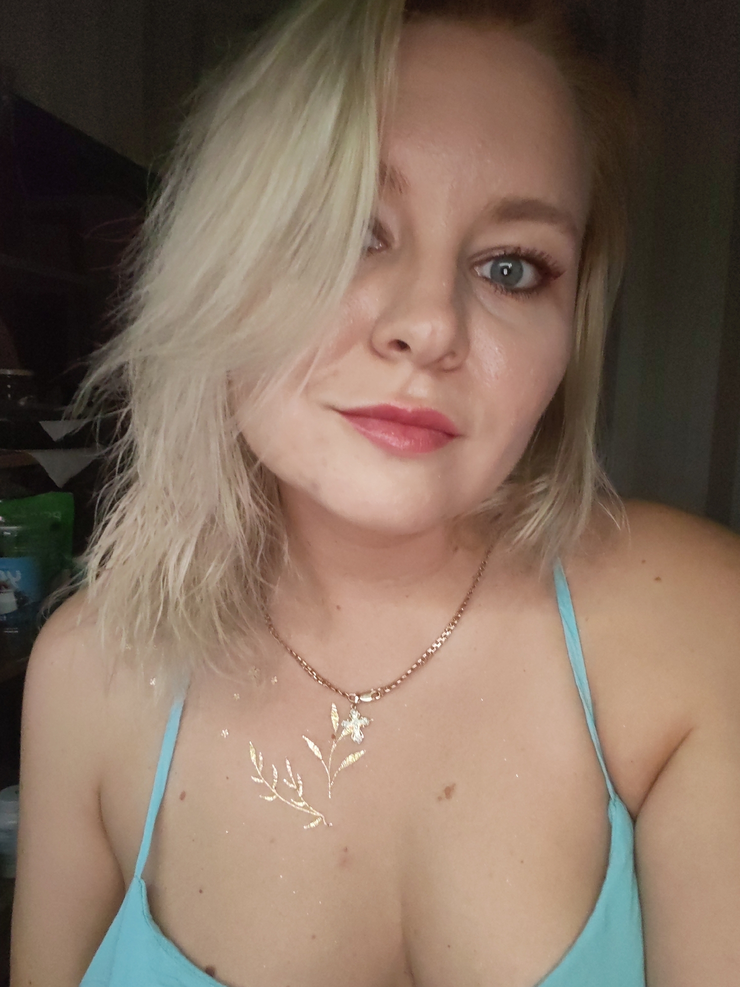 The image is a close-up, head-and-shoulders shot of a young woman with blonde, messy hair and light blue eyes. She is wearing a light blue top and a rose gold chain necklace with a floral pendant. Her chest is adorned with a temporary gold glitter tattoo of a floral design. The background is dark and out of focus. The image was taken in Izhevsk, Russia.