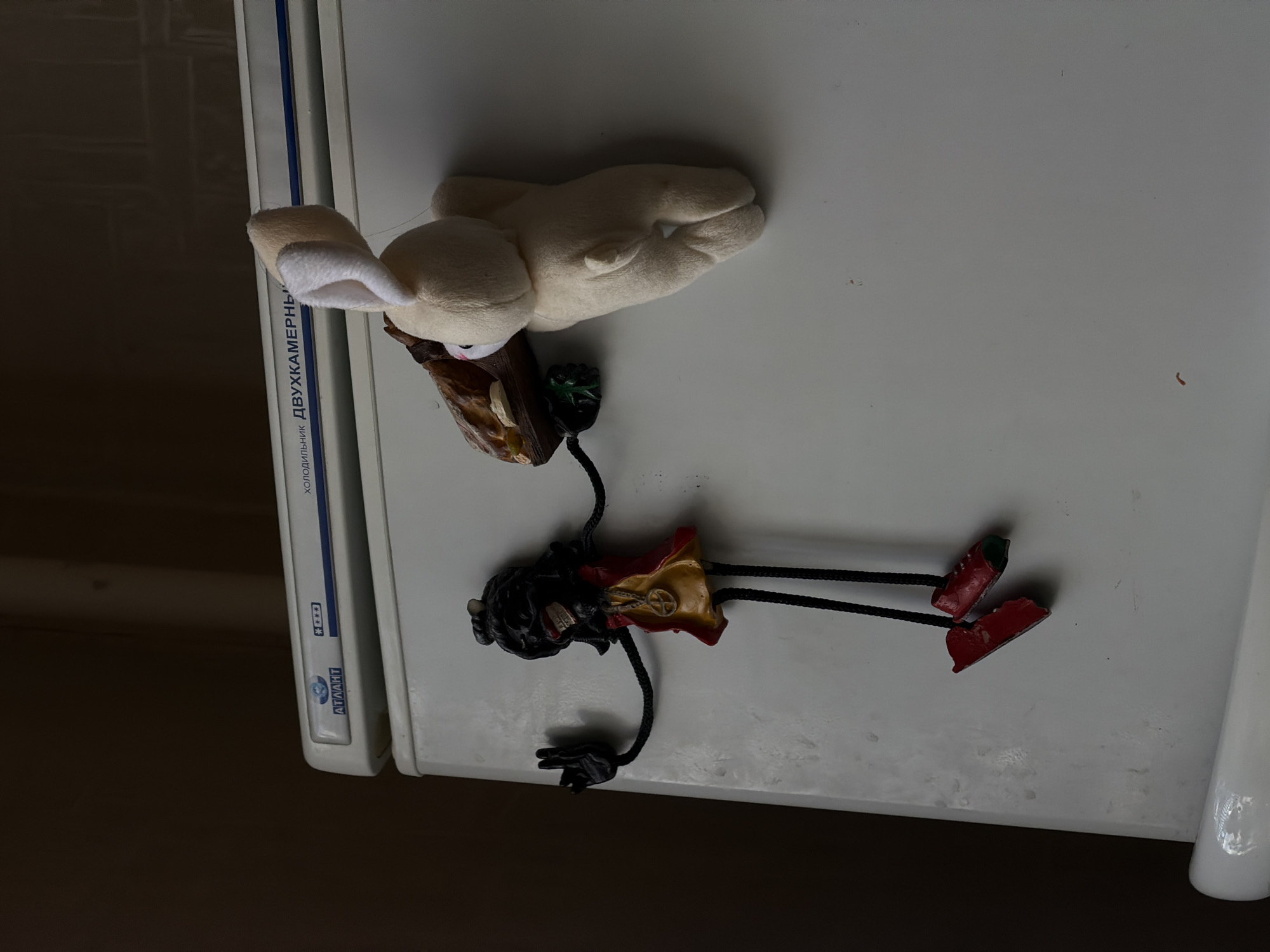 The image, taken from a top-down perspective, displays two decorative items resting on the white, glossy top surface of an appliance, likely a refrigerator. The image is rotated 90 degrees clockwise.
On the left, a cream-colored plush toy, possibly a rabbit or fennec fox, lies on its back. It has long, plush ears with white inner linings and small, dark eyes. The toy appears to be holding a small, irregularly shaped brown object, which might be a piece of chocolate or a decorative wooden item, adorned with a few green leaves.
Below and slightly to the right of the plush toy is a distinctive figurine. This character features a dark, stylized head with an open mouth revealing teeth. It wears a brightly colored outfit, predominantly red with yellow sections, and a circular emblem, possibly a peace sign, on its chest. Its limbs are thin, black, cord-like structures, ending in small black hands and distinct red, boot-like shoes. The figurine appears to be suspended or lying beside the plush toy.
The appliance beneath these items is white and features a blue and black stripe along its side. Visible text in Cyrillic script includes "ХОЛОДИЛЬНИК ДВУХКАМЕРНЫЙ" (Kholodil'nik Dvukhkamernyy), meaning "Two-compartment Refrigerator," and the brand name "ATЛАНТ" (ATLANT). The background to the left of the appliance is a dark, indistinct brown, suggesting a shadowed wall or furniture. The items are stationary, and no specific activity or event is taking place. No particular time of day or weather conditions can be inferred from the lighting.