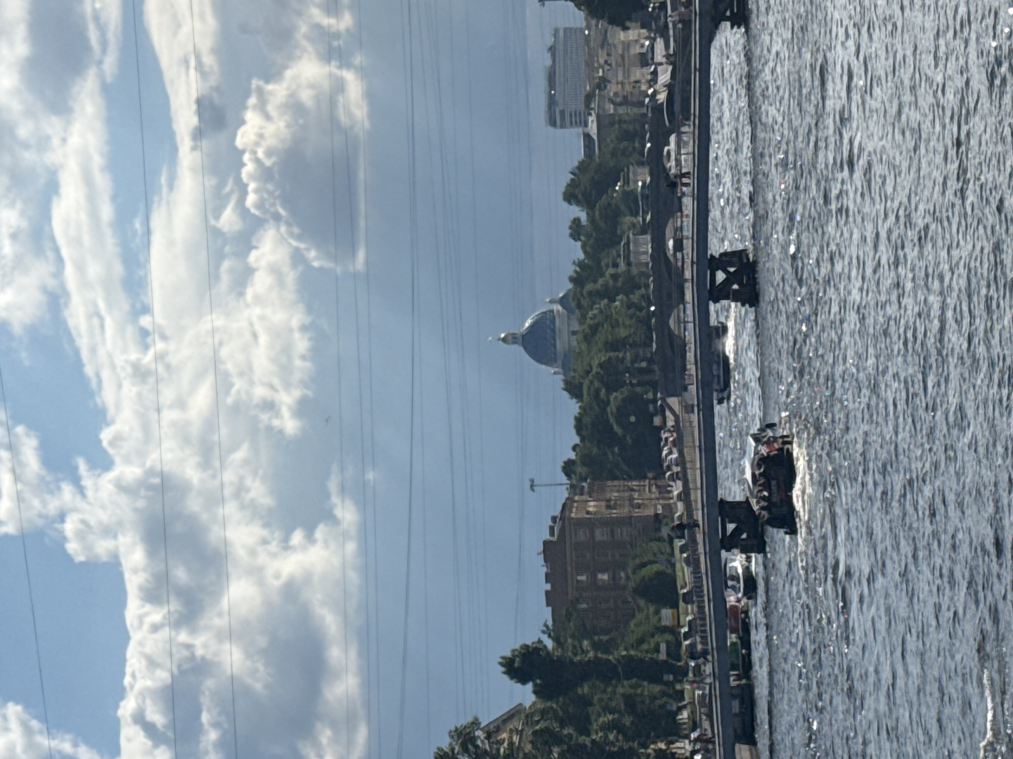 The image depicts a wide river in Saint Petersburg, Russia, under a partly cloudy sky. In the foreground, the river's surface is choppy with sunlight glinting off the water. Several boats are navigating the river. One boat, in the middle ground, is a dark-colored, flat-topped vessel with people visible on its deck. Further back, a larger bridge spans the river, with a white boat passing underneath it. On the far bank, a cityscape unfolds, featuring trees, buildings, and a prominent blue-domed building that appears to be a cathedral or significant architectural landmark. The architecture suggests a historical European city. Overhead, power lines are strung across the sky, partially obscuring the clouds. The overall impression is a sunny day with moderate river traffic in a scenic urban setting.