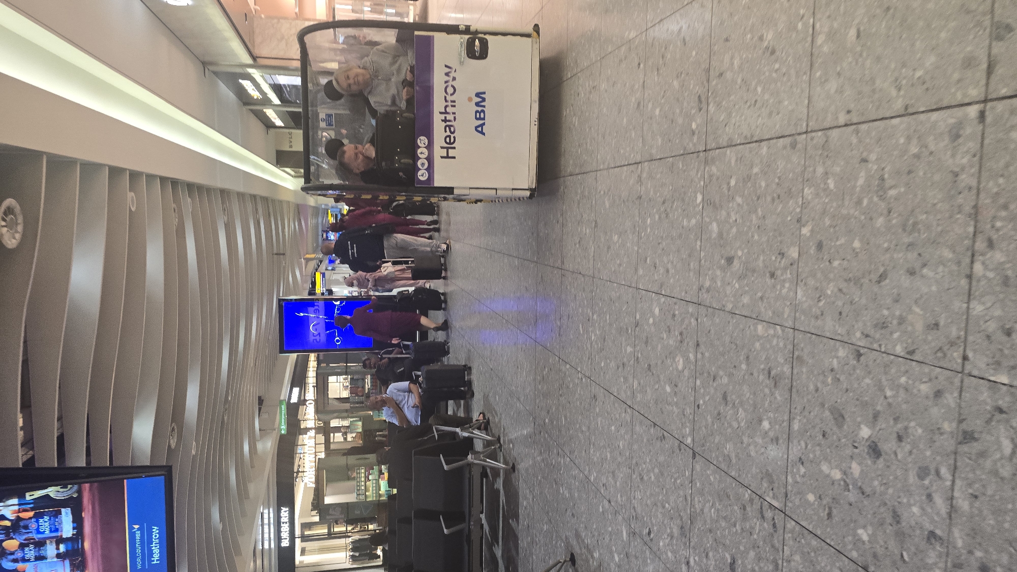 The image shows a scene inside Heathrow Airport, London, United Kingdom. In the foreground, there is a motorized vehicle with "Heathrow ABM" written on its side, carrying two passengers. Further along, a group of people are walking with luggage. In the middle ground, there is a seating area with people waiting, and a Burberry store. A digital display screen is visible. The floor is covered with grey tiles, and the ceiling has a curved design.